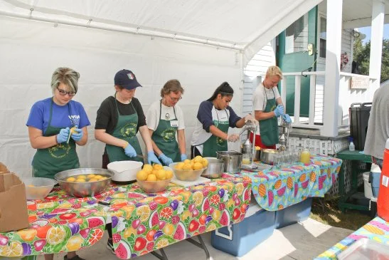 Five people preparing food at a colorful outdoor table with lemons and kitchen utensils during a sunny day.