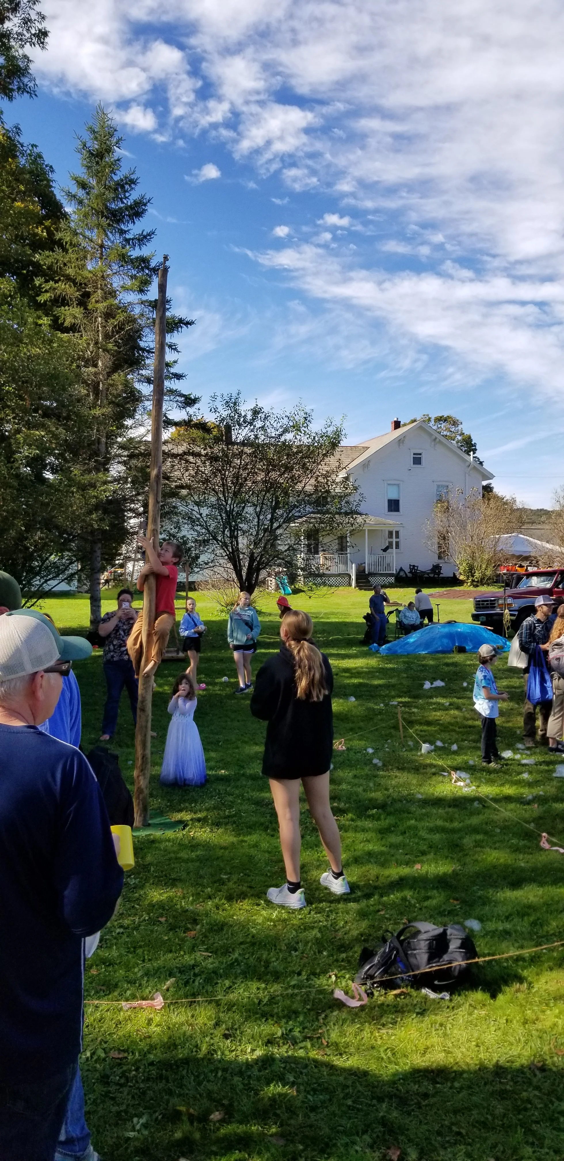 Children and adults at an outdoor event, with a girl climbing a pole to reach a prize. People are standing on grass, some holding papers or looking at the activity. A house and trees are visible in the background, with a blue sky and scattered clouds overhead.