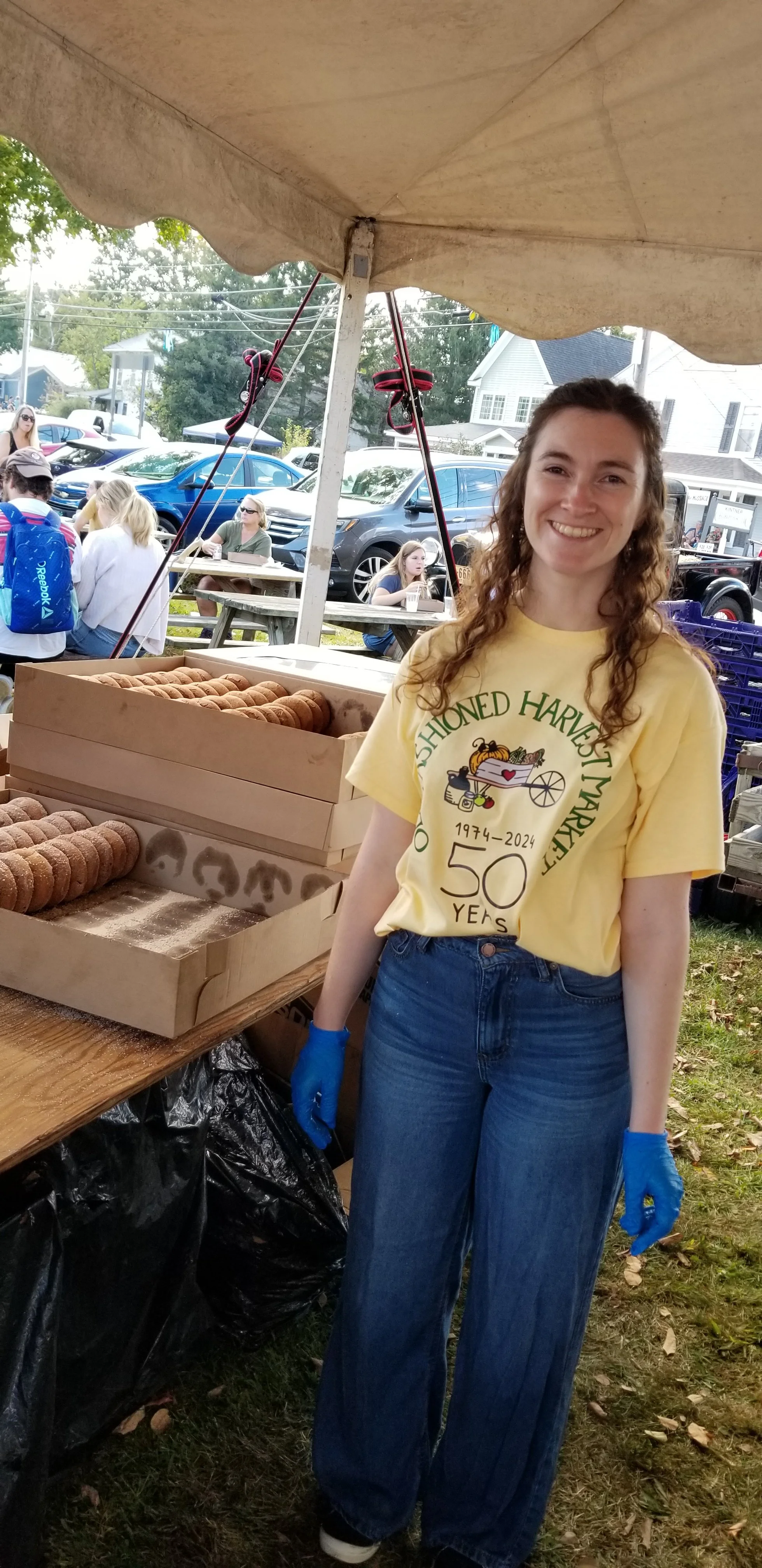 A young woman wearing a yellow T-shirt and blue jeans standing next to a table with trays of cookies at an outdoor event.