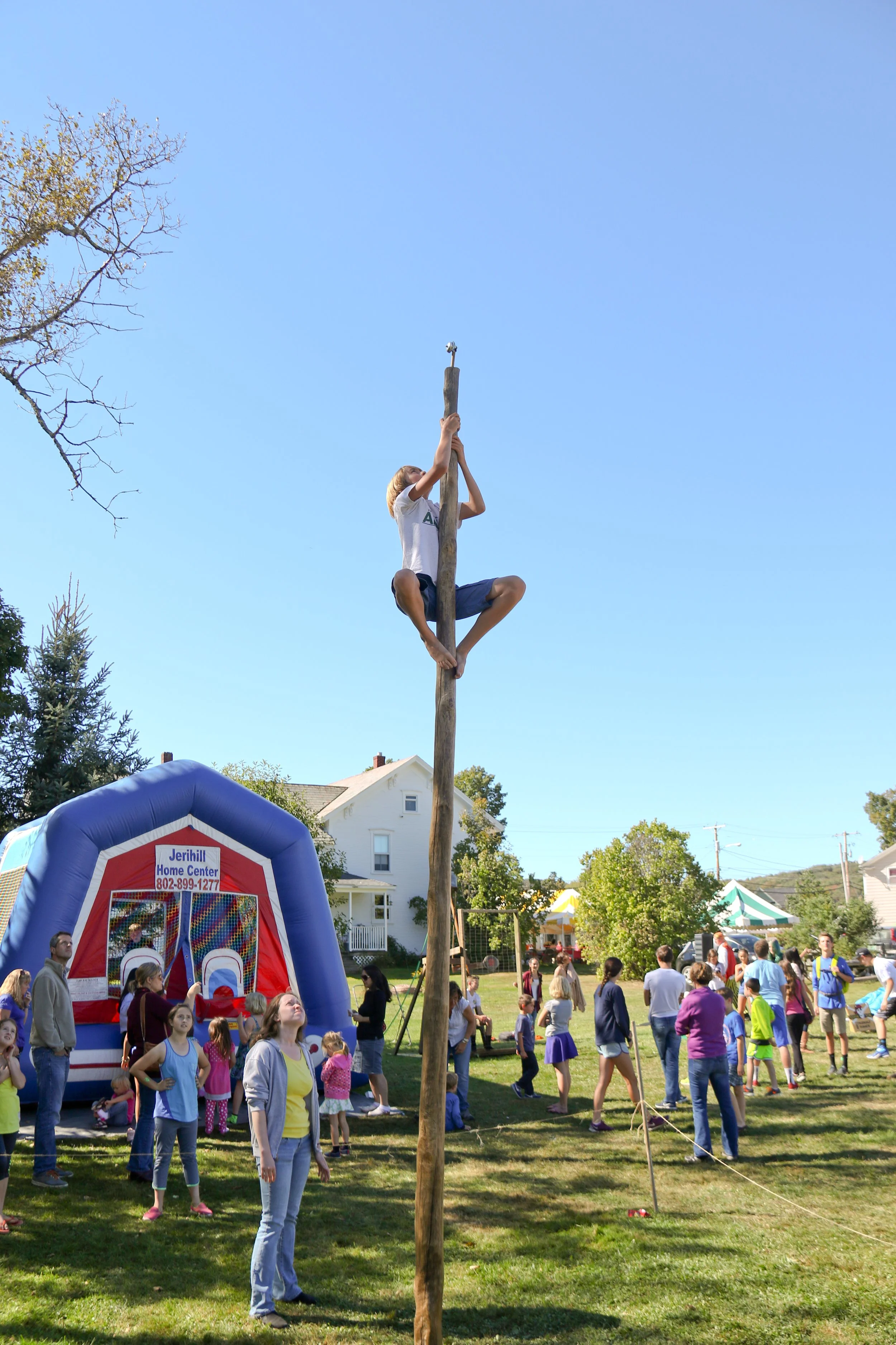 A person climbing a tall wooden pole during an outdoor community event, with many people watching and participating in various activities.