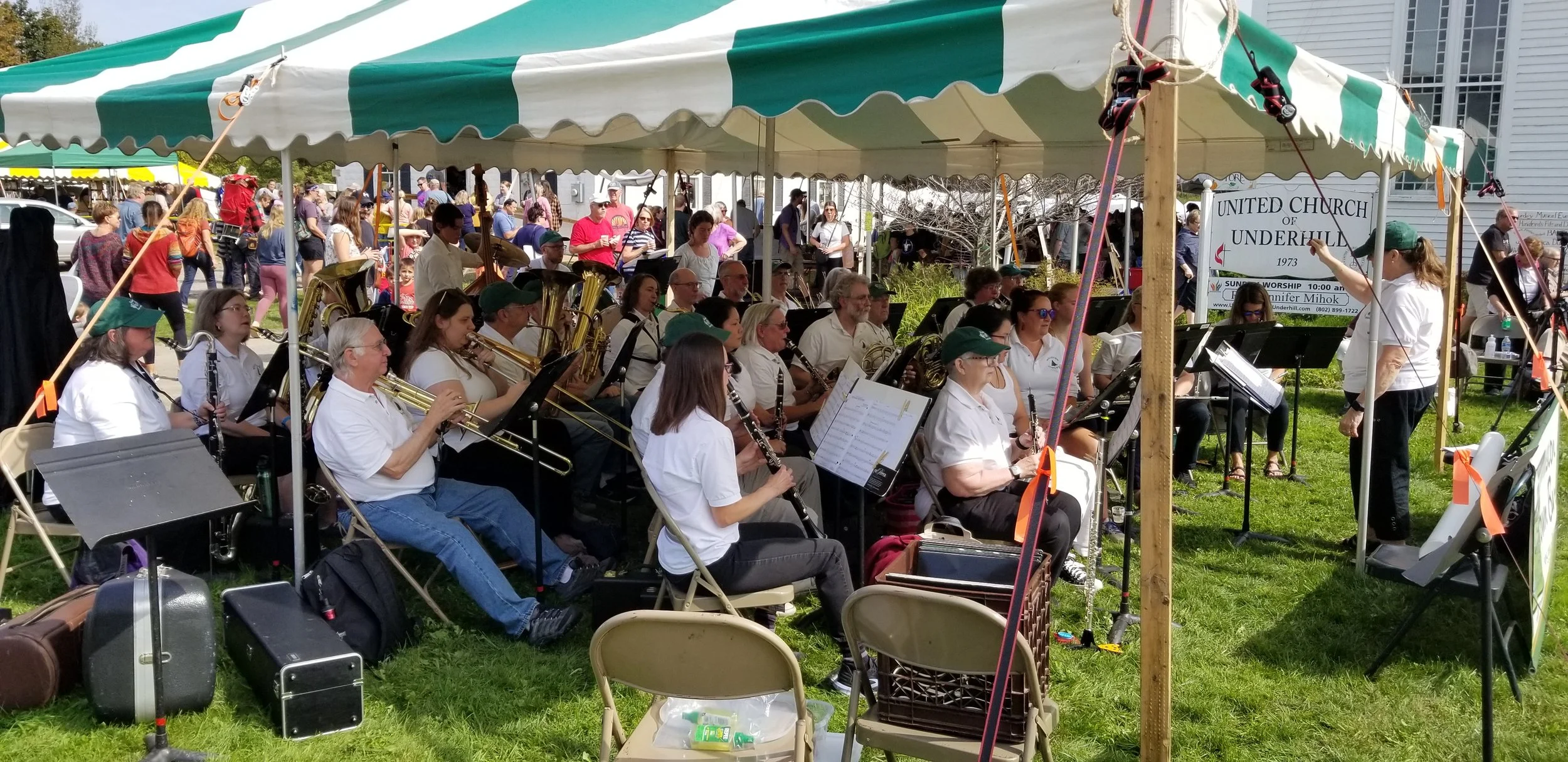 Outdoor music at the Harvest Market, featuring a large band or orchestra under a canopy tent, with many musicians playing various instruments such as trumpets, clarinets, and saxophones.