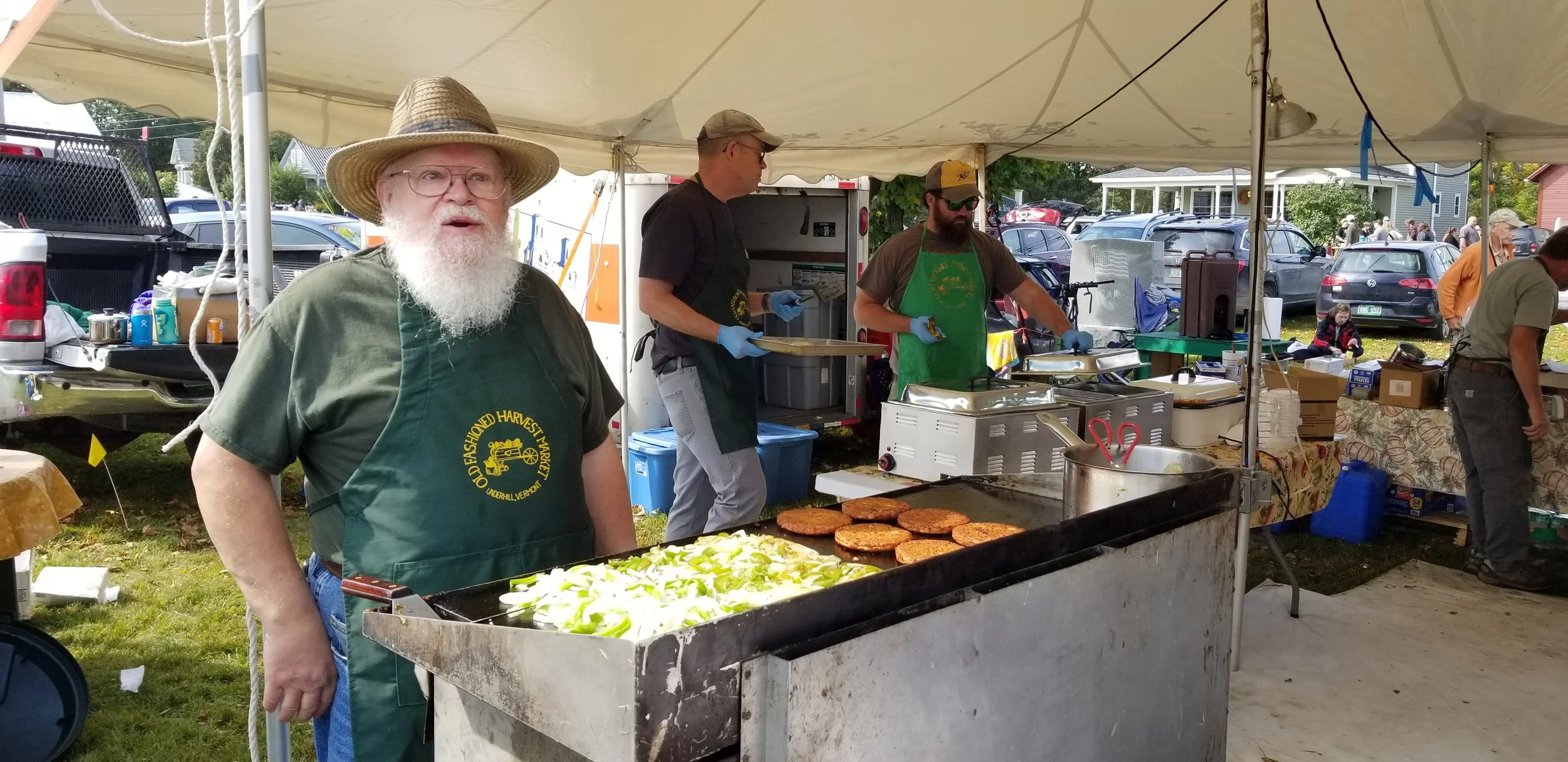 Man with a beard and glasses wearing a straw hat and green apron standing at a food stand with cooked patties and chopped vegetables, preparing food at an outdoor market.