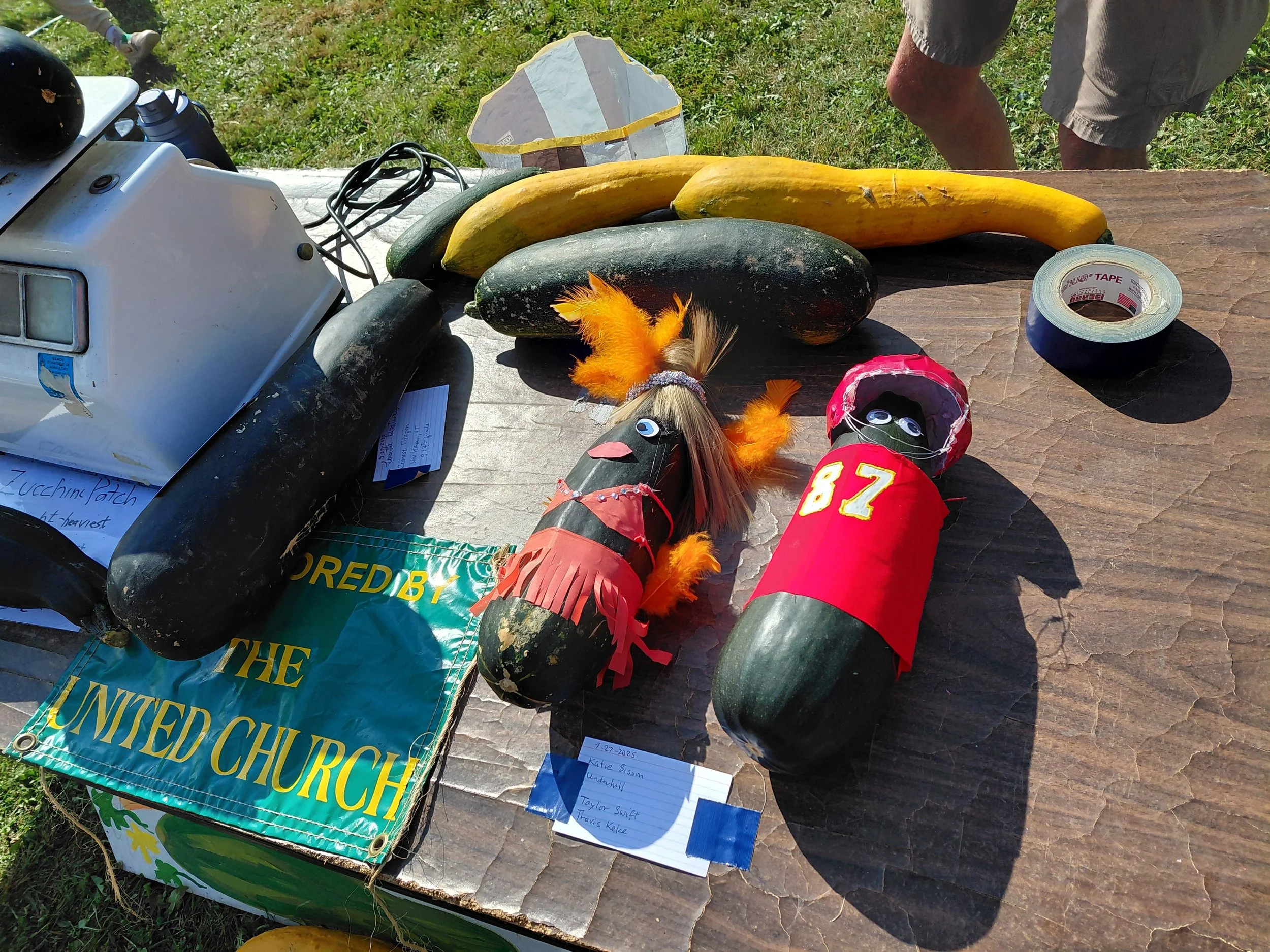 Display table with pumpkins, decorative scarecrows, a booklet, and church signs at an outdoor event.