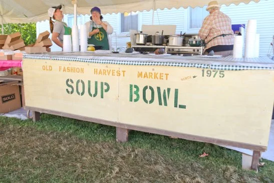 People at a soup booth at a fall festival, with a sign reading 'Old Fashion Harvest Market, Soup Bowl, Since 1975' and tables filled with soup pots and cups.