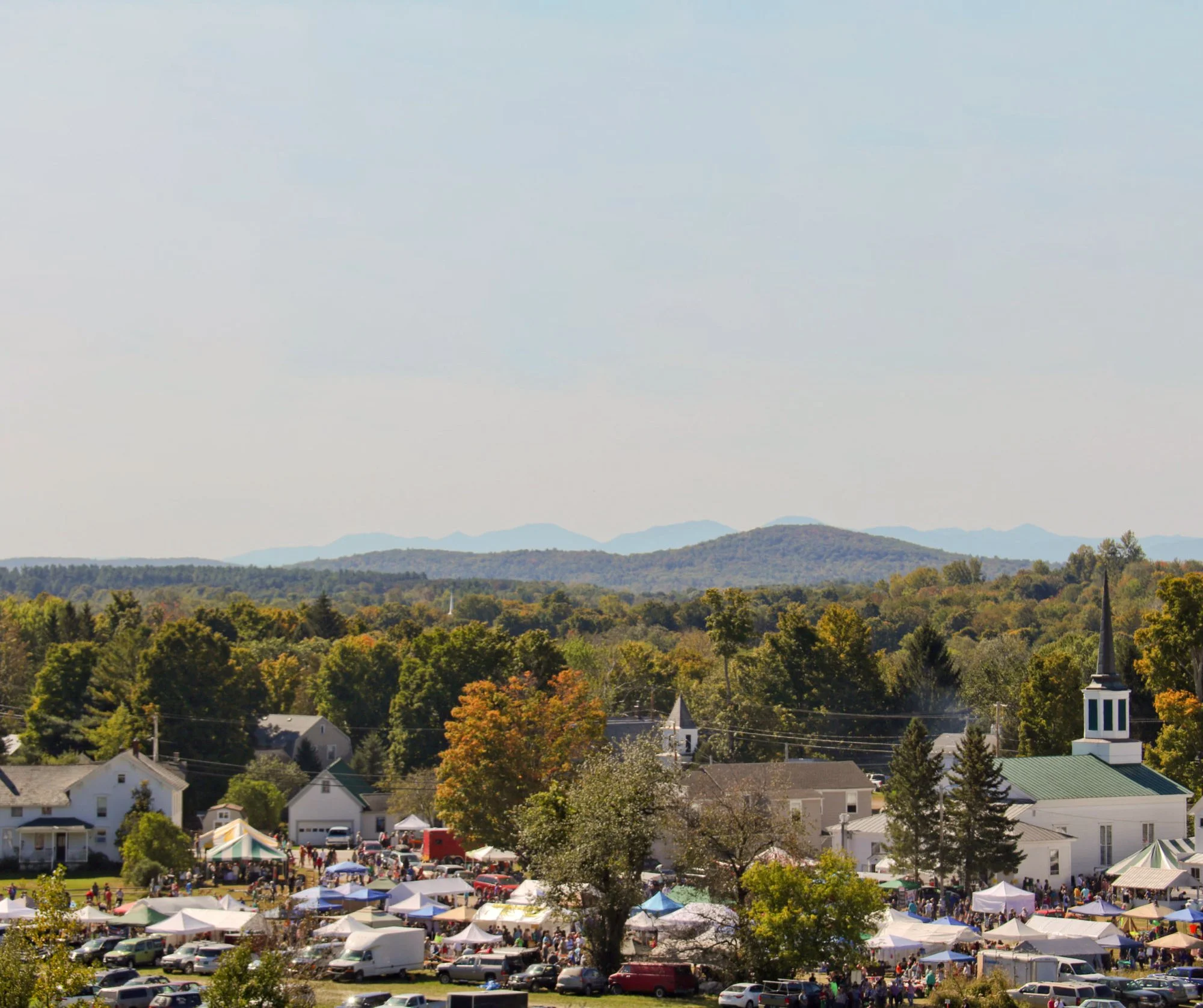 A small town's outdoor event with tents, stalls, and many people, surrounded by lush green trees and distant hills under a cloudy sky.