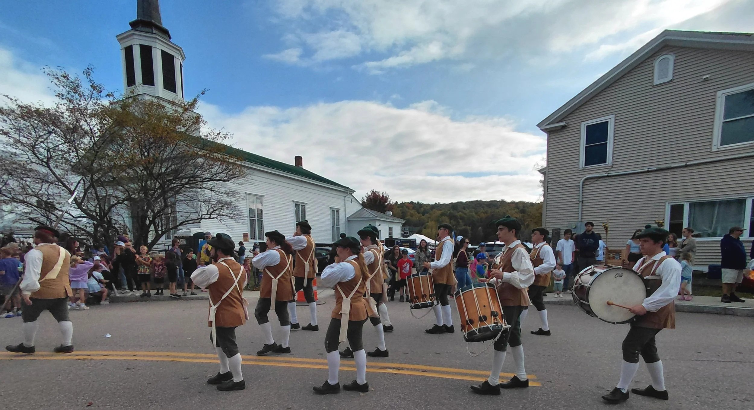 A parade with a marching band in vintage-style uniforms playing drums and other instruments as spectators watch from the sidewalk in a small town with a church and houses in the background.