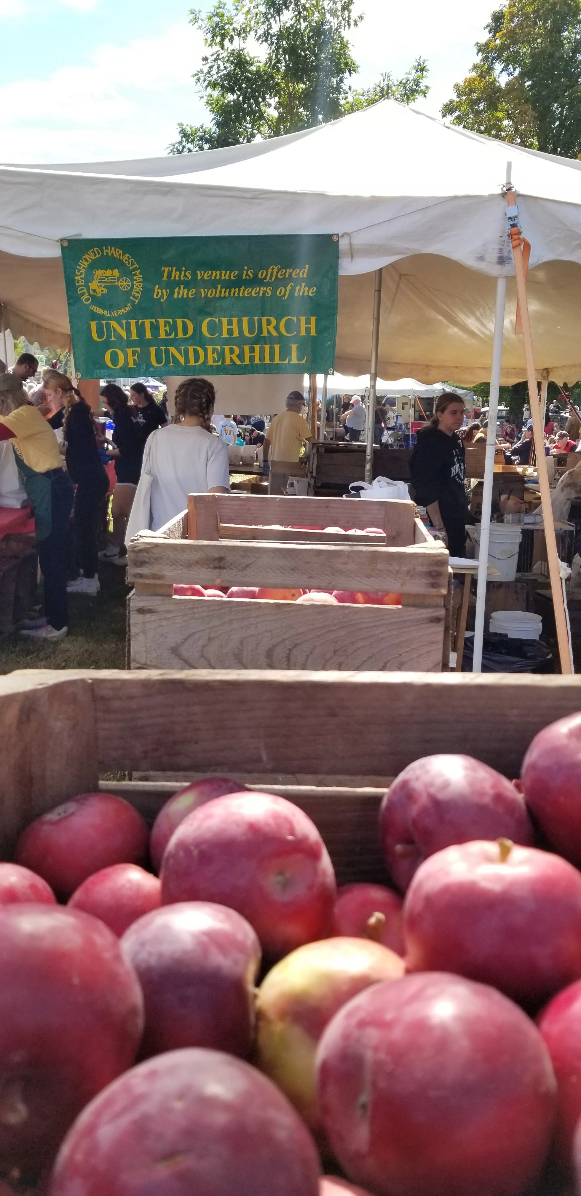 A group of people at an outdoor market under a white tent, with a sign indicating the venue is offered by the volunteers of the United Church of Underhill. The foreground shows a wooden crate filled with red apples.