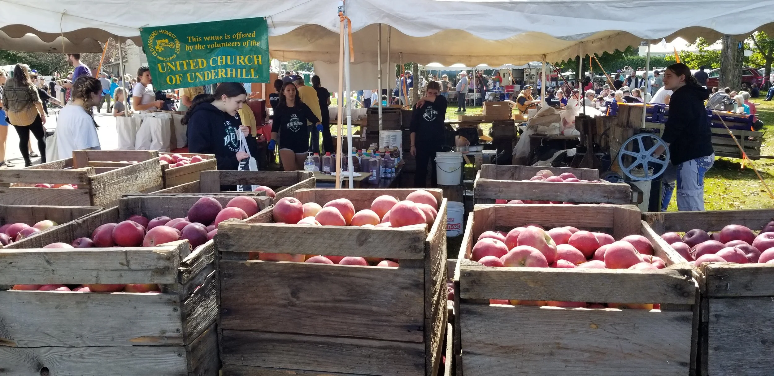 A gathering of people at an outdoor farmers market with wooden bins filled with pink and red apples, under a canopy with a banner reading 'United Church of Underhill'.