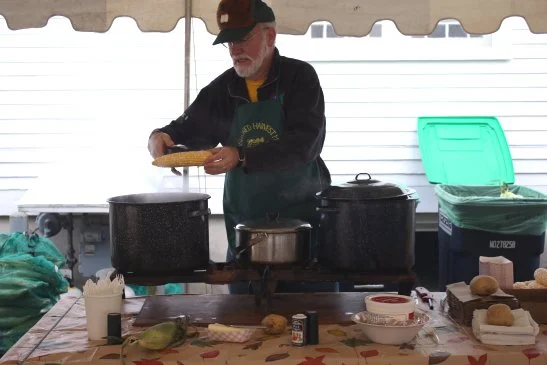 Man cooking at an outdoor food stall with large pots, a cutting board, and various food items on the table.