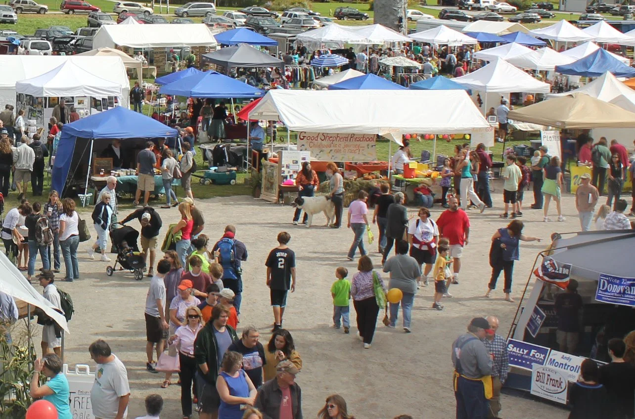 People walking and browsing the flea market with numerous stalls and tents, including a food stand for Good Shepherd Preschool, a political campaign booth, and vendors selling various items on a grassy field.