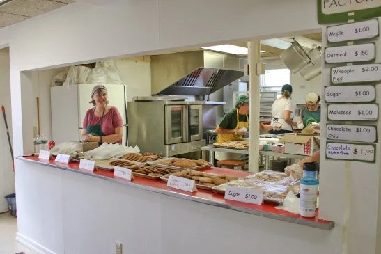 Staff behind a bakery counter with baked goods and a menu of prices, with two employees in background preparing food.