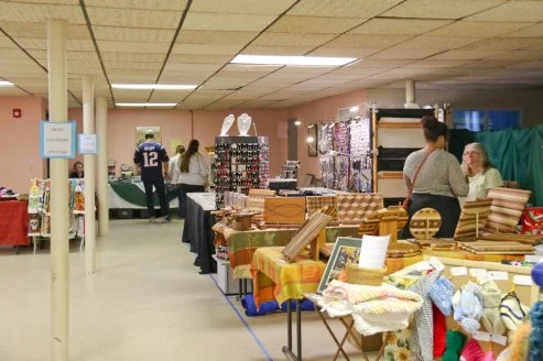 Indoor craft fair with tables displaying handmade textiles, jewelry, and accessories, and people browsing and shopping.