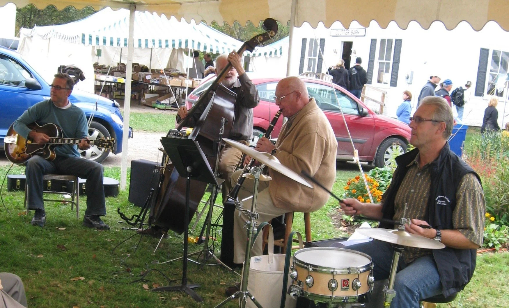 Four musicians performing outdoors under a tent, playing guitar, upright bass, clarinet, and drums.