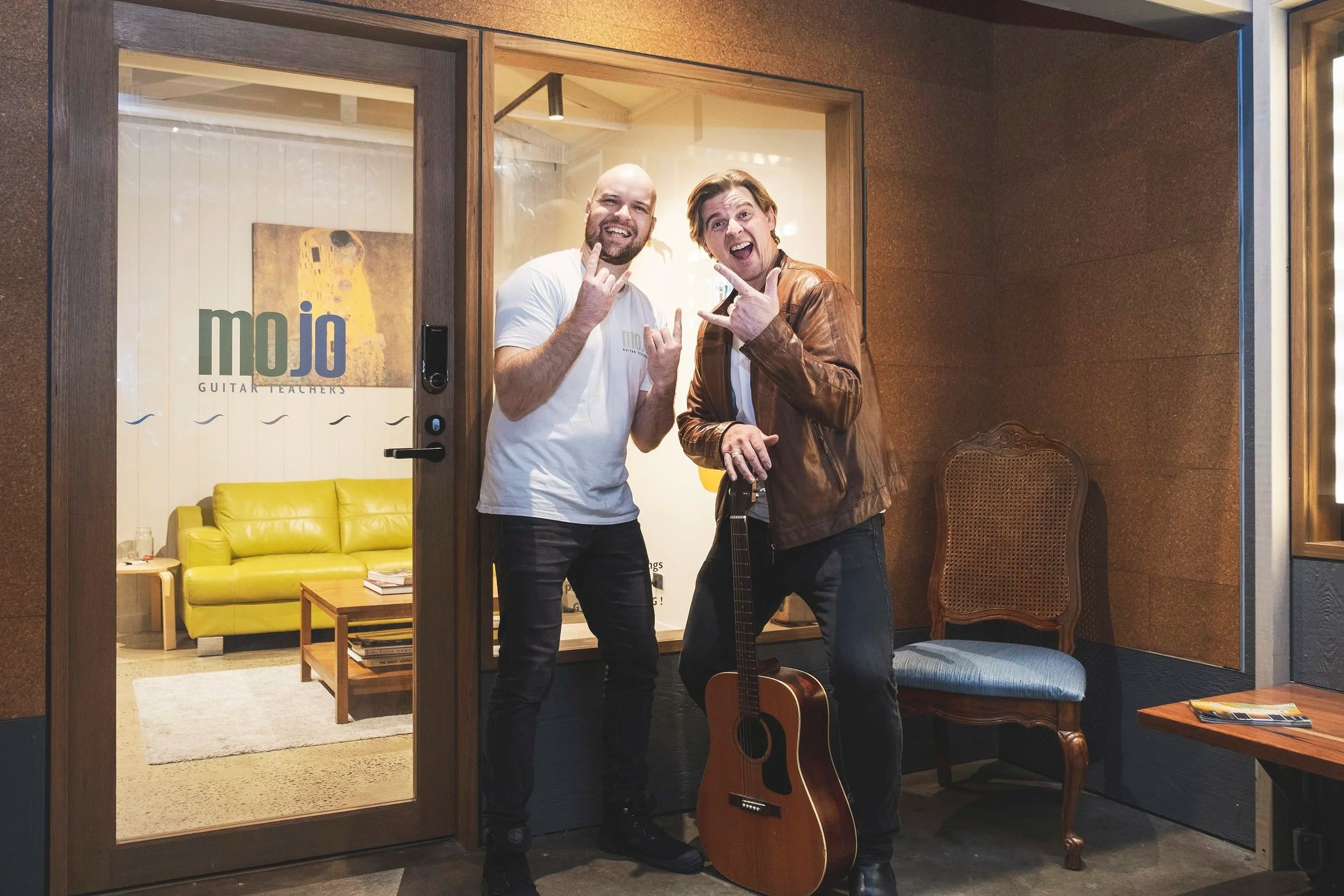 Cam & Dan the founders of MojoDojo, one holding a guitar, both making rock hand gestures, inside a glass-walled room with a yellow sofa and coffee table in the background.