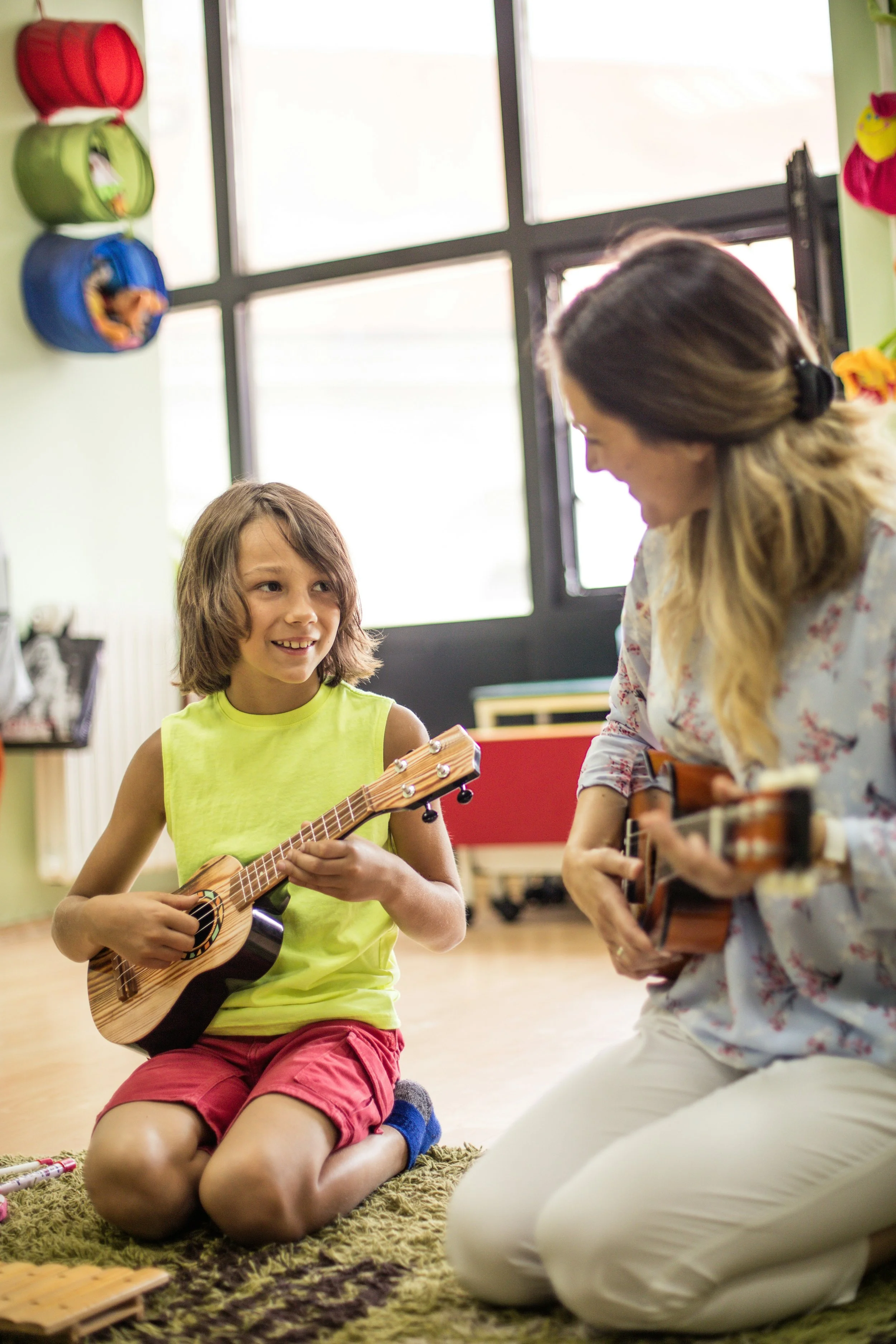 A woman and a young girl playing guitars together in a bright room with colorful decorations and large windows.