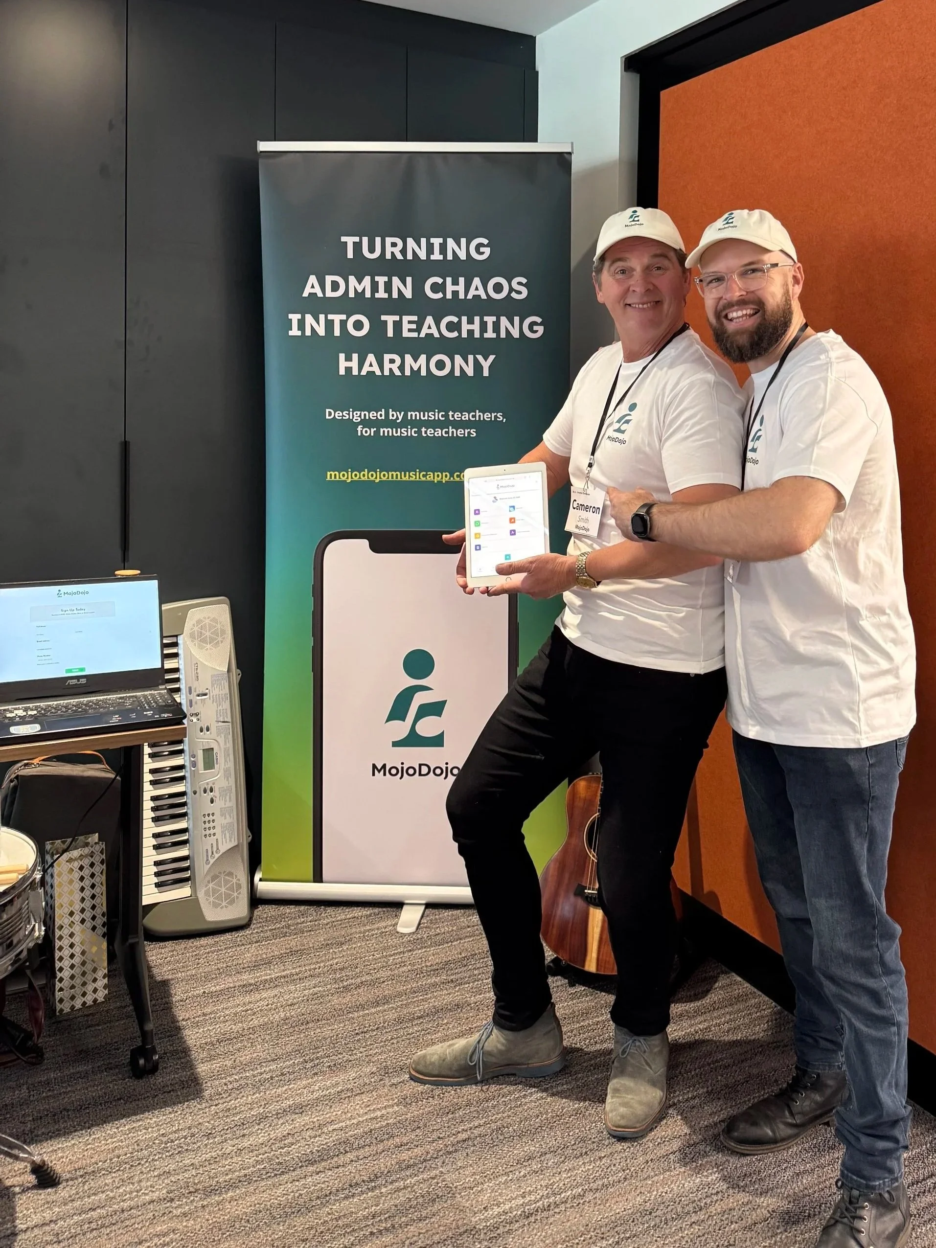 Two men smiling and posing with a tablet in front of a MojoDojo banner at a music education event. The banner reads 'Turning admin chaos into teaching harmony' and mentions music teachers. One man is wearing a white cap and a white MojoDojo t-shirt, holding the tablet, while the other man is wearing glasses, a white MojoDojo t-shirt, and has his arm around the first man. There is a keyboard and a laptop on a table nearby.