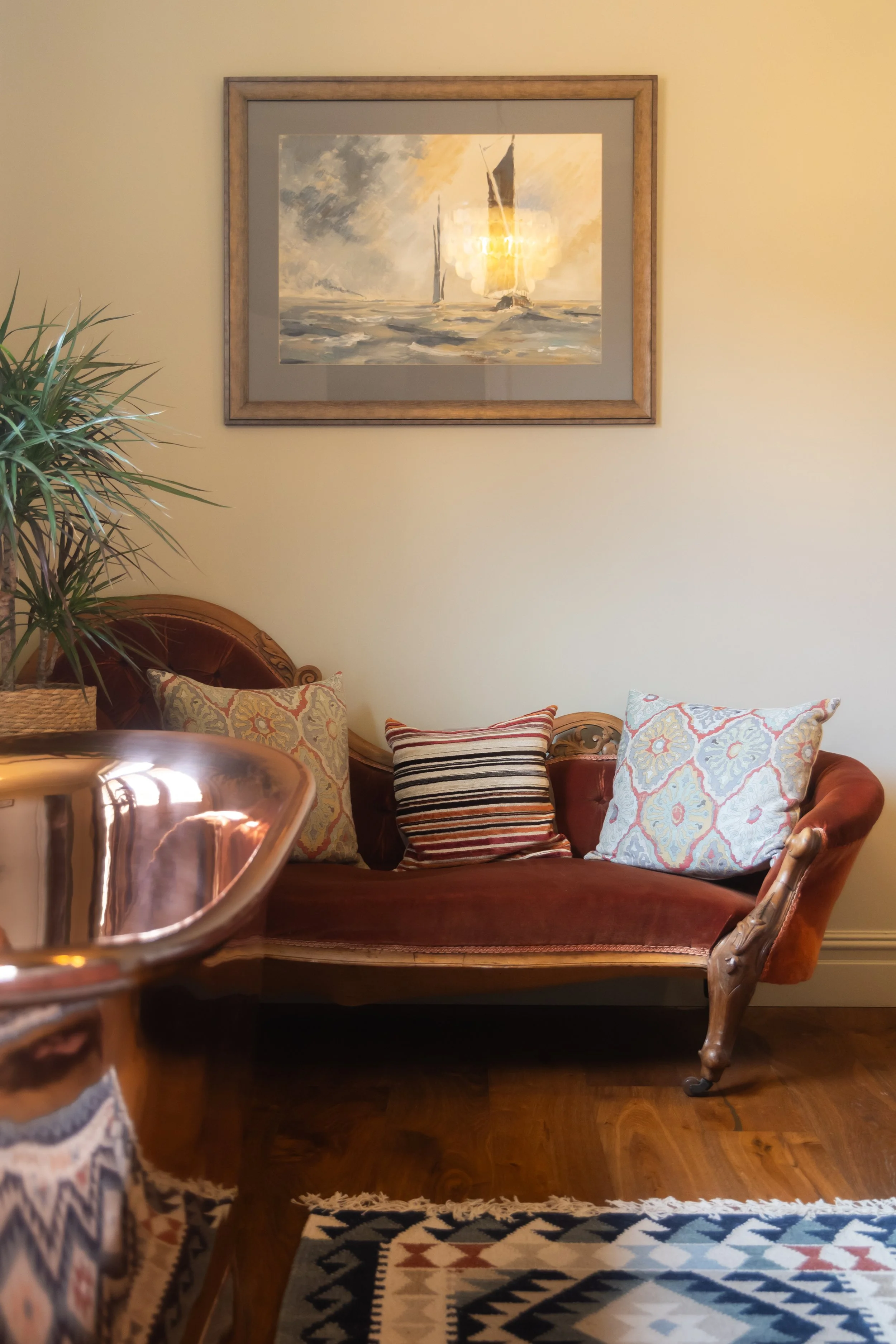 Living room with a vintage red velvet sofa, patterned pillows, a potted plant, a wooden side table, a patterned rug, and a painting of sailboats on the wall.