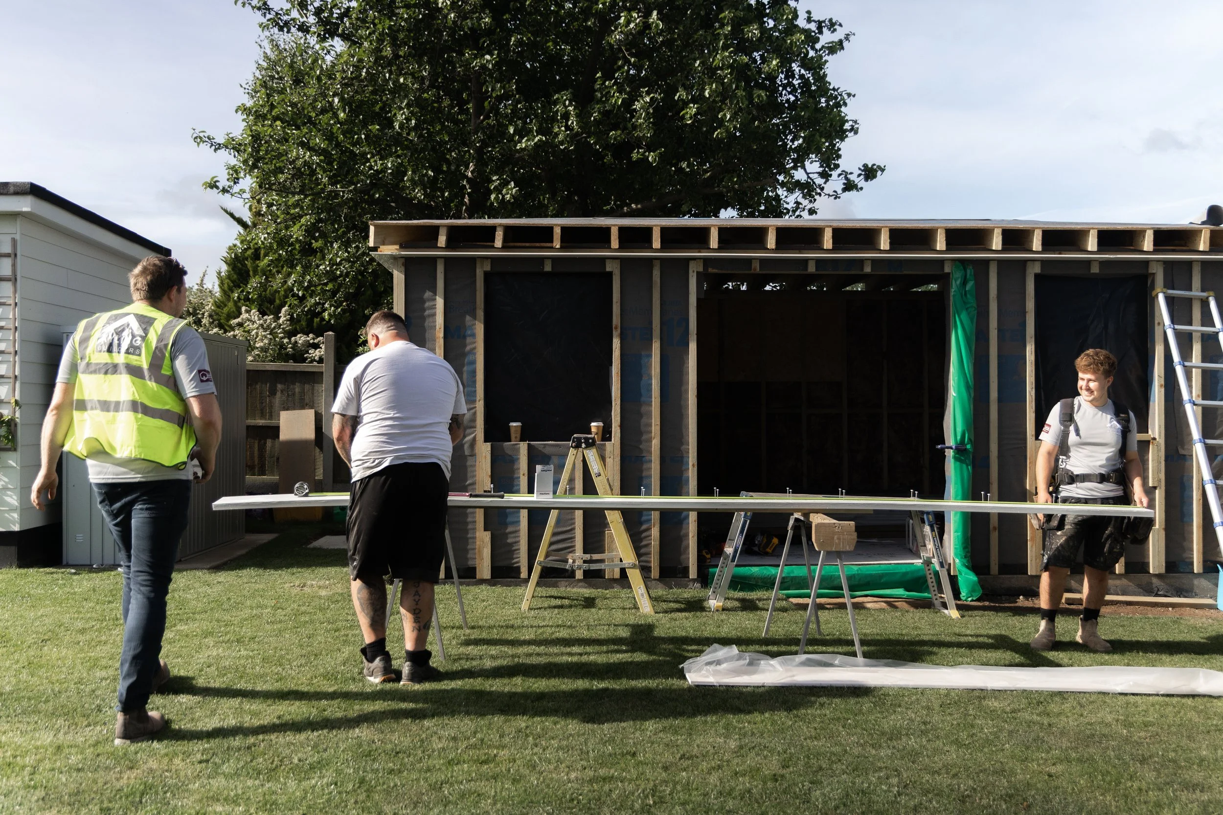 Four men are working on constructing a small wooden shed in a backyard, with a large tree and a partly cloudy sky in the background.