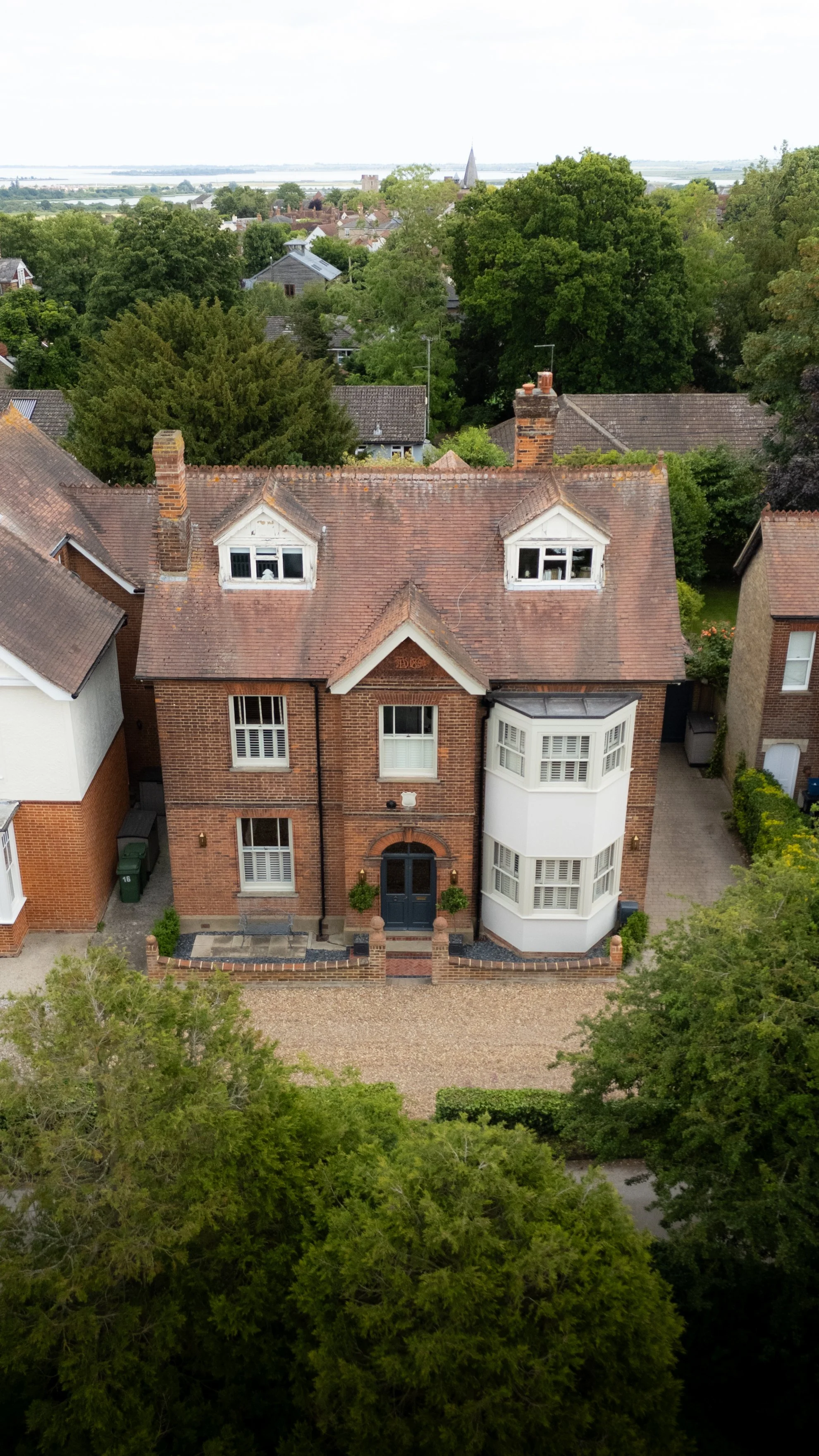 An aerial view of a red-brick house with a turret and large bay window, surrounded by green trees, with other houses in the background and a body of water in the distance.