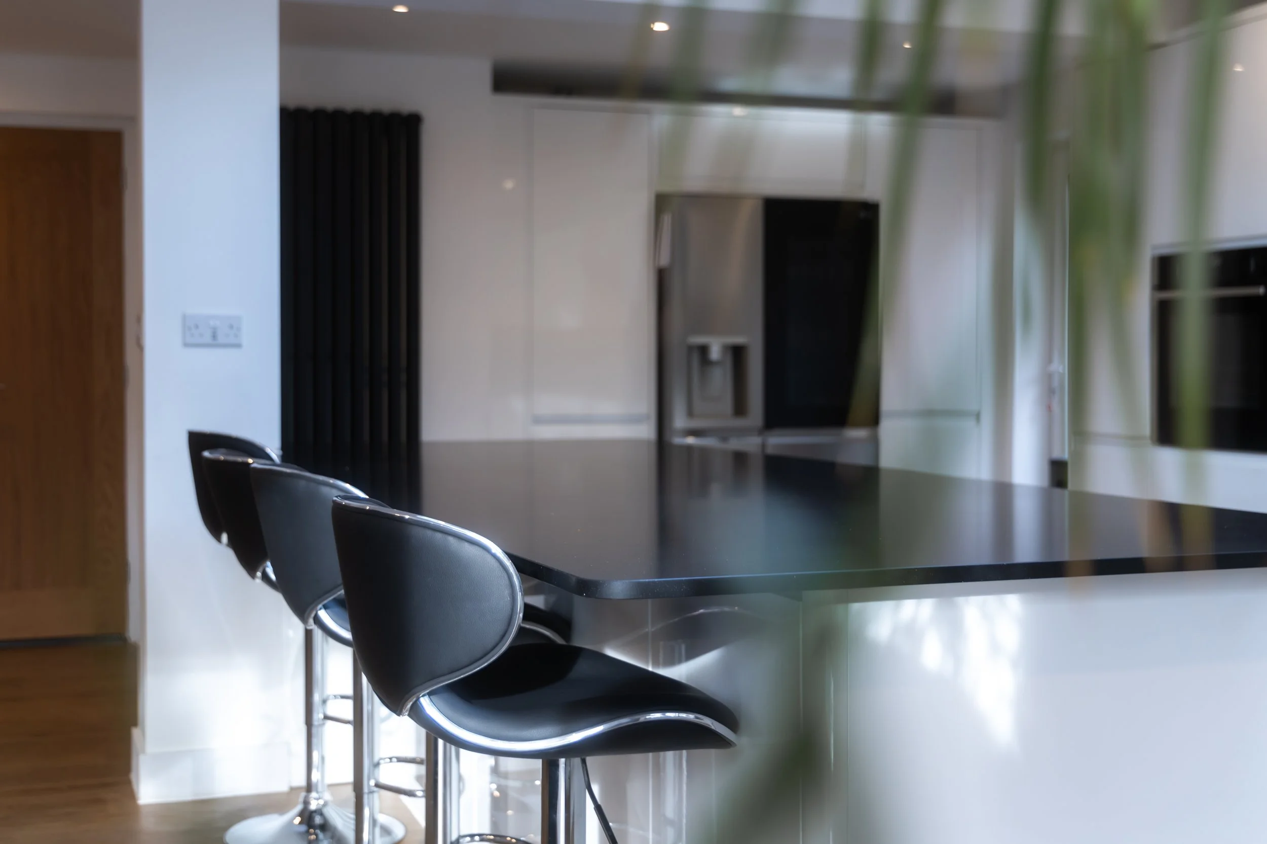 Interior view of a modern kitchen with a black countertop island, three black chairs at the island, white cabinets, built-in stainless steel oven, and a window in the background.