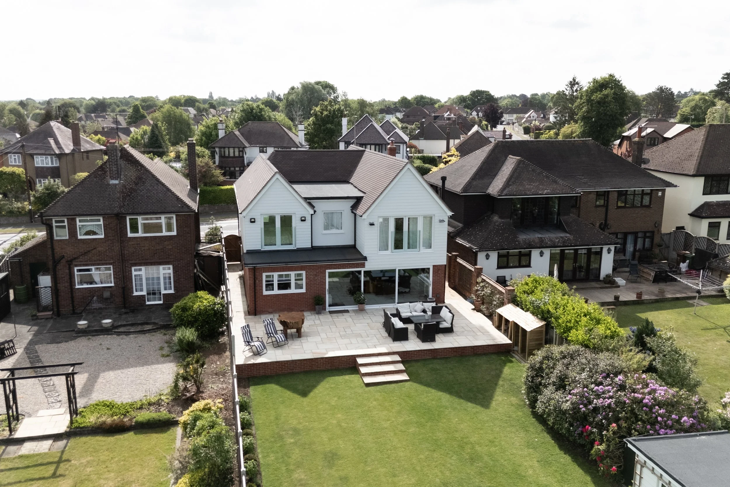 Aerial view of a suburban neighborhood with multiple houses, a backyard patio with outdoor furniture, lush green lawn, and trees.