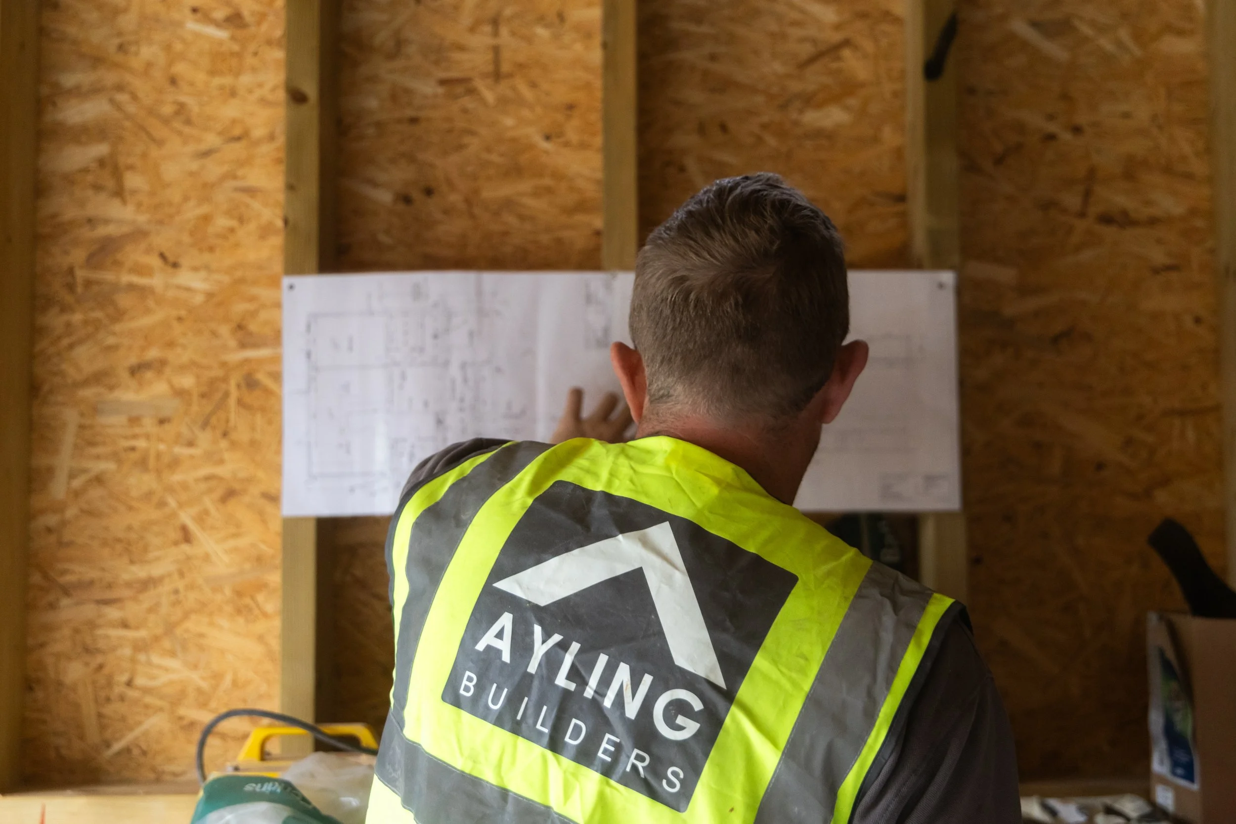 Construction worker with short hair wearing a high-visibility vest with the words "AVIL BLDG" visible on the back, working inside a wooden structure.