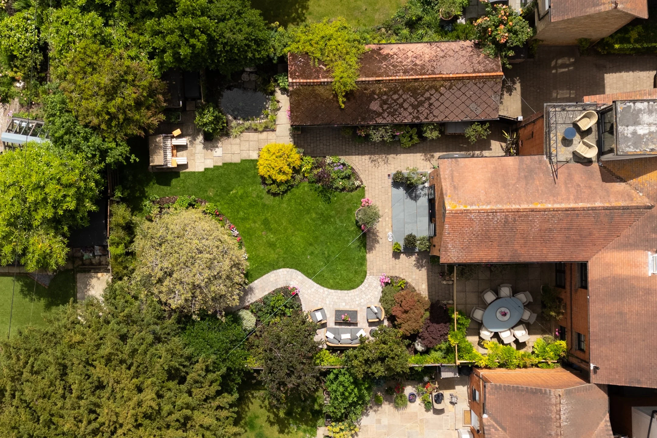 Aerial view of a backyard with lush green trees, a brick house with a red roof, a stone pathway, outdoor furniture, and a grassy lawn.