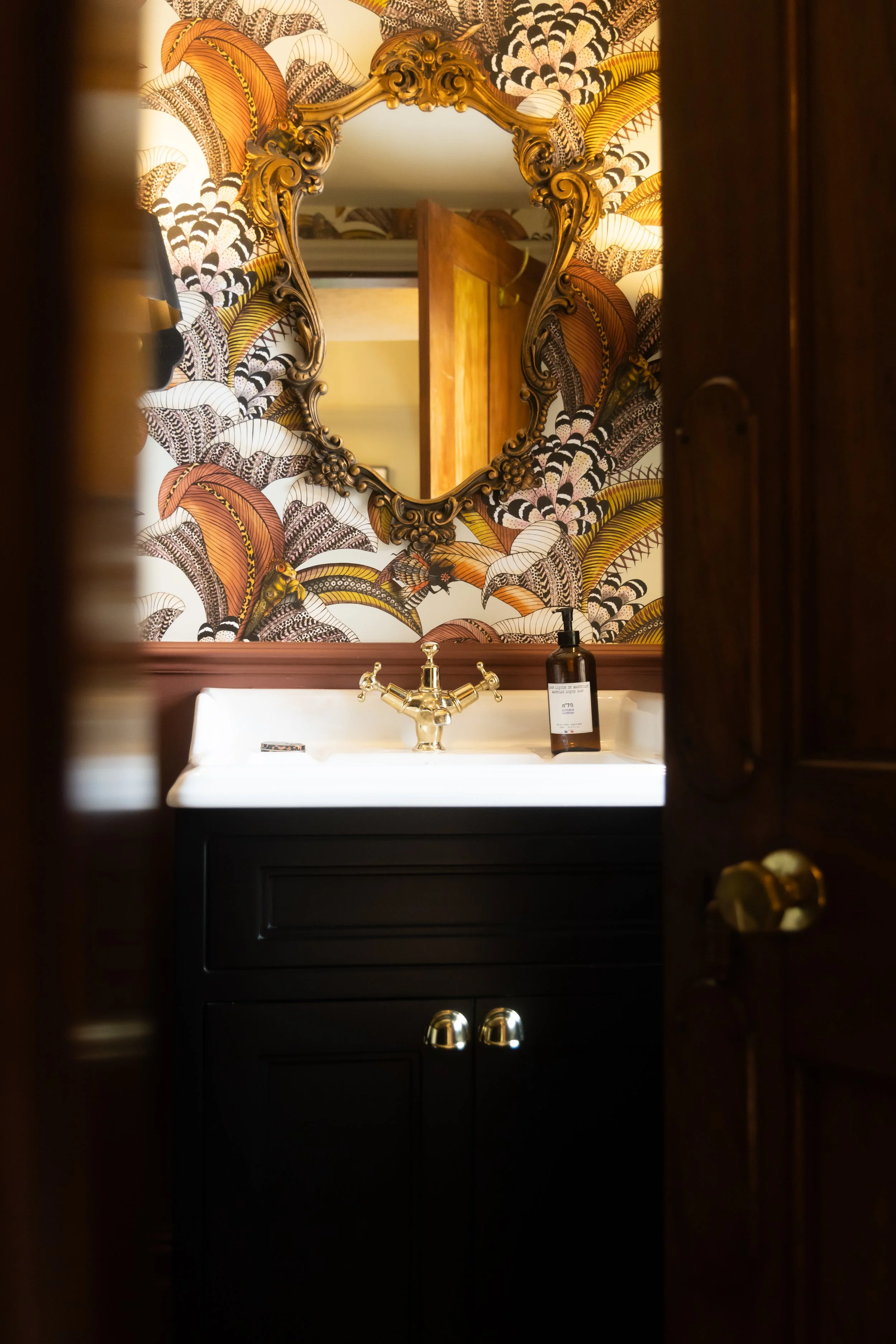 A mirror with a gold ornate frame hanging on a wallpapered wall featuring colorful abstract feather designs in shades of yellow, brown, black, and white. Below the mirror, a white sink with a gold faucet is visible, with a brown soap dispenser and a 