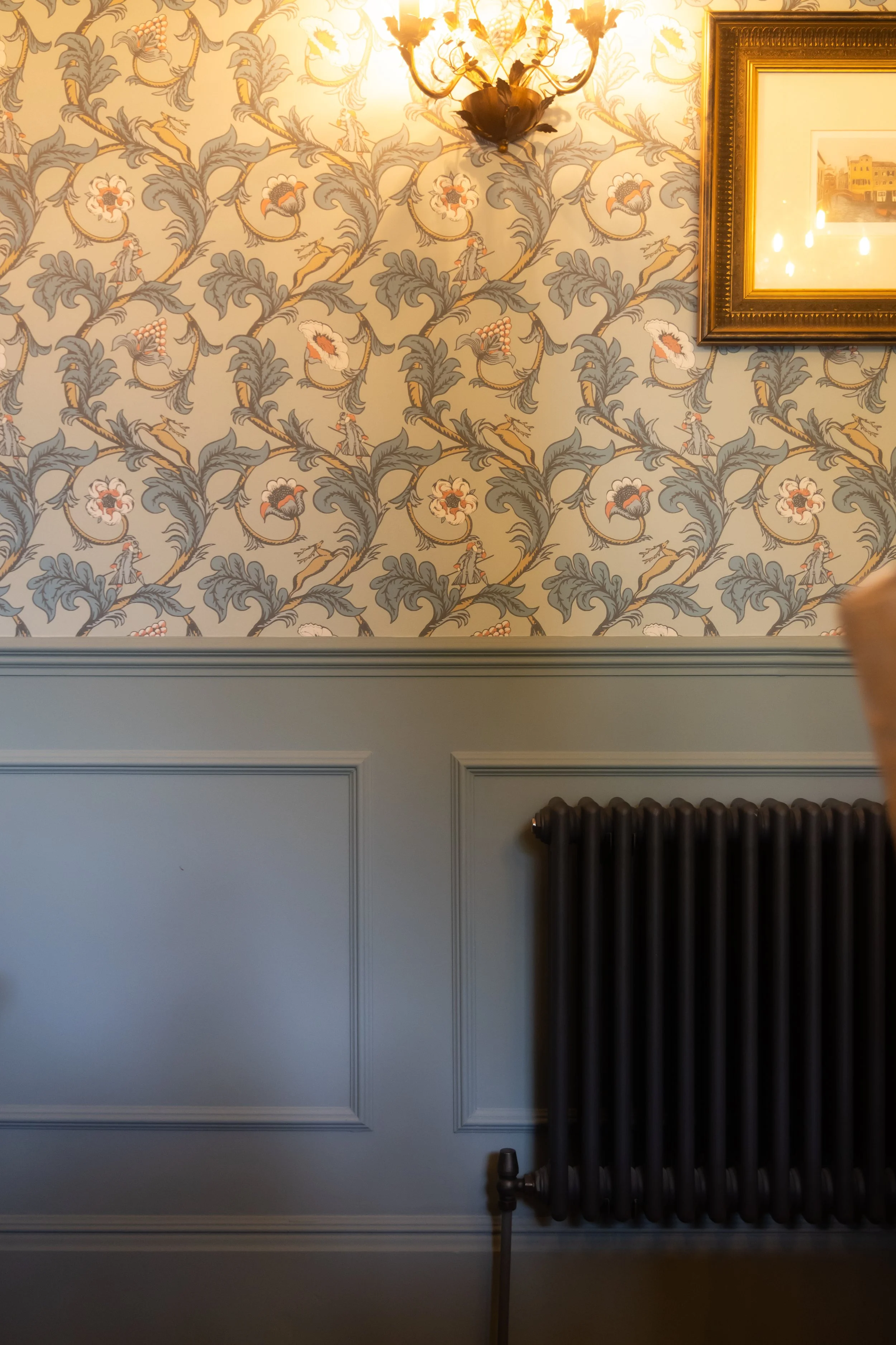 A wall with ornate floral wallpaper, a framed painting, a wall-mounted light fixture resembling a chandelier, and a black radiator in the corner.