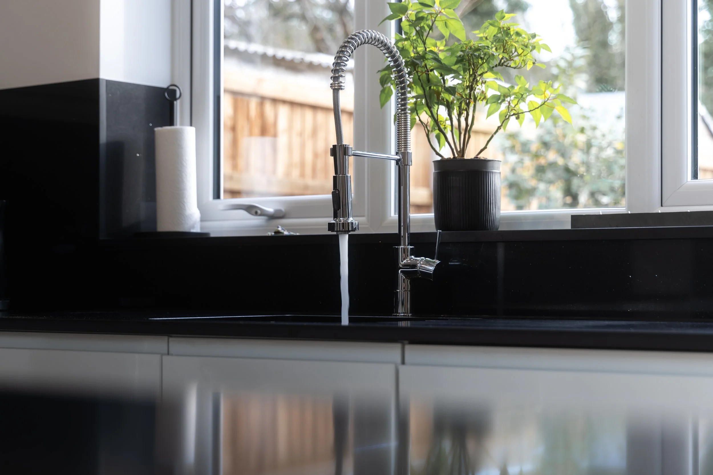 Close-up of kitchen sink with modern chrome faucet and green potted plant on window sill, outside view of a wooden fence and trees.