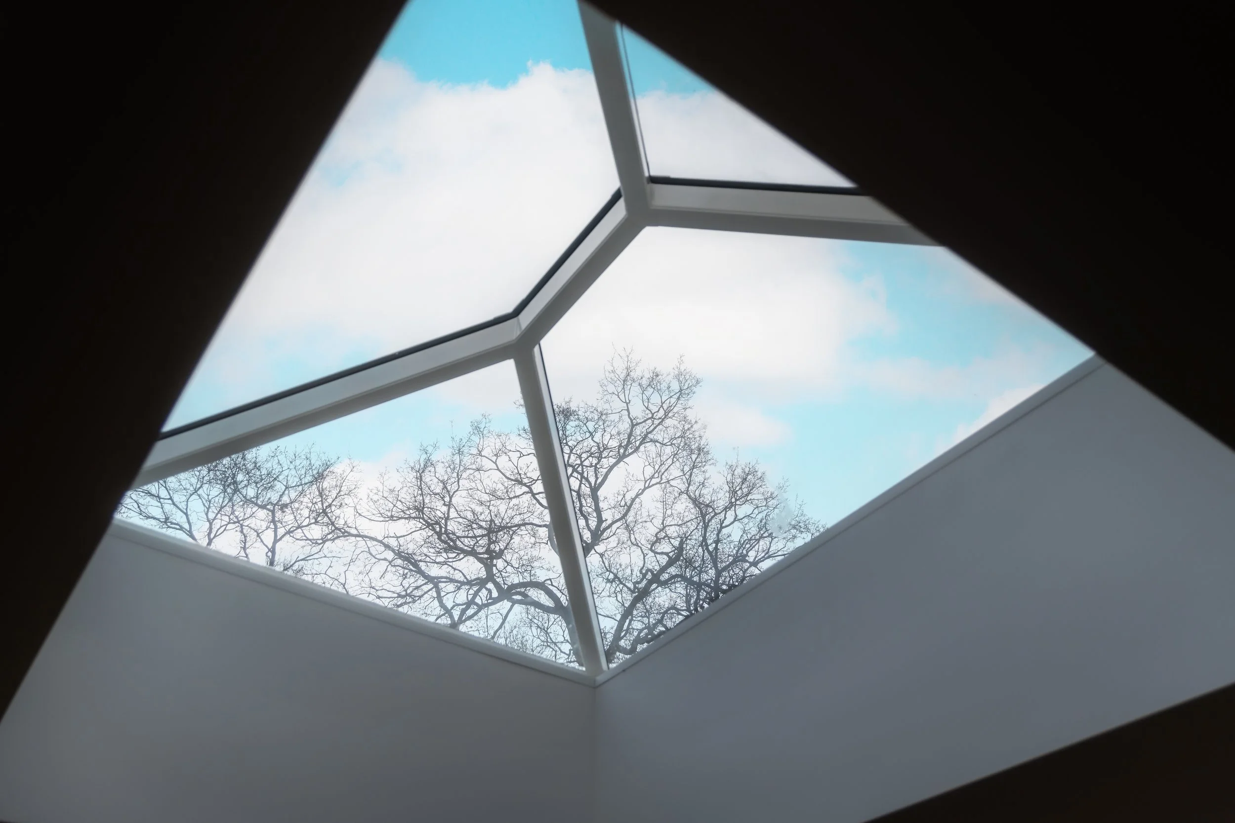 Looking up through a skylight or roof window at a cloudy sky with blue patches and leafless trees outside.