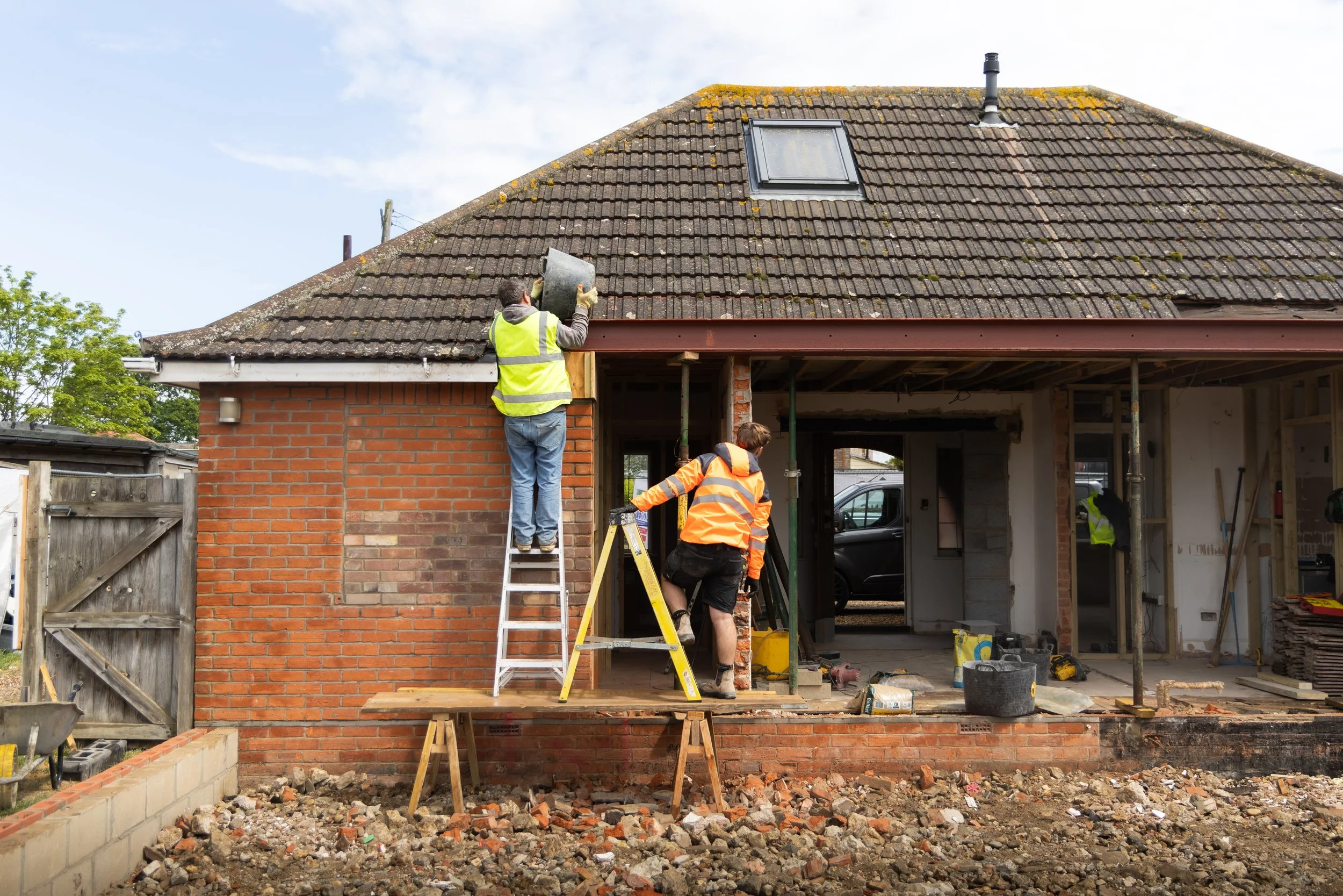 Construction workers repair a house roof with new tiles, while some work on ladders and scaffolding on the house's brick exterior. The work area is filled with tools, debris, and construction materials.