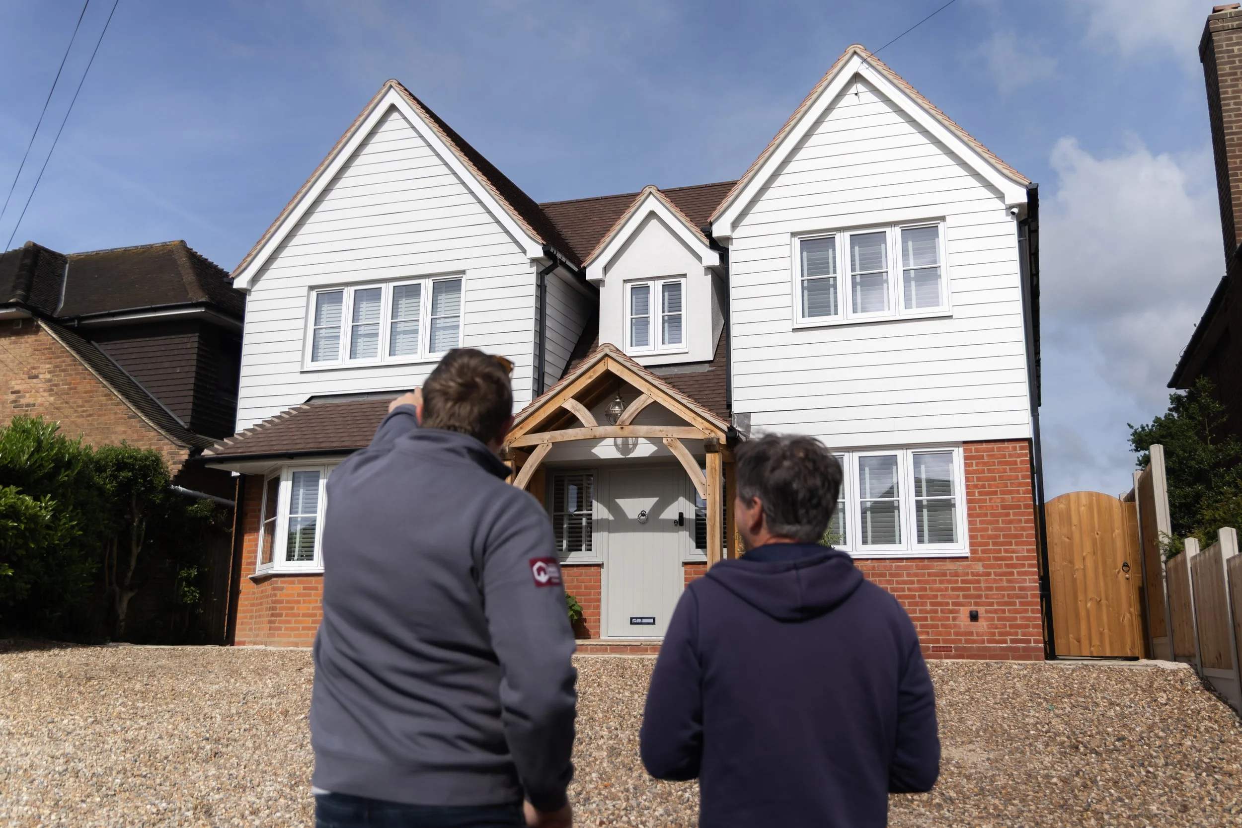 Two men standing in front of a white, two-story house with gabled roof, dark shingles, and multiple windows. One man is pointing towards the house, while the other looks on. The house has a wooden entrance canopy and a gravel driveway.