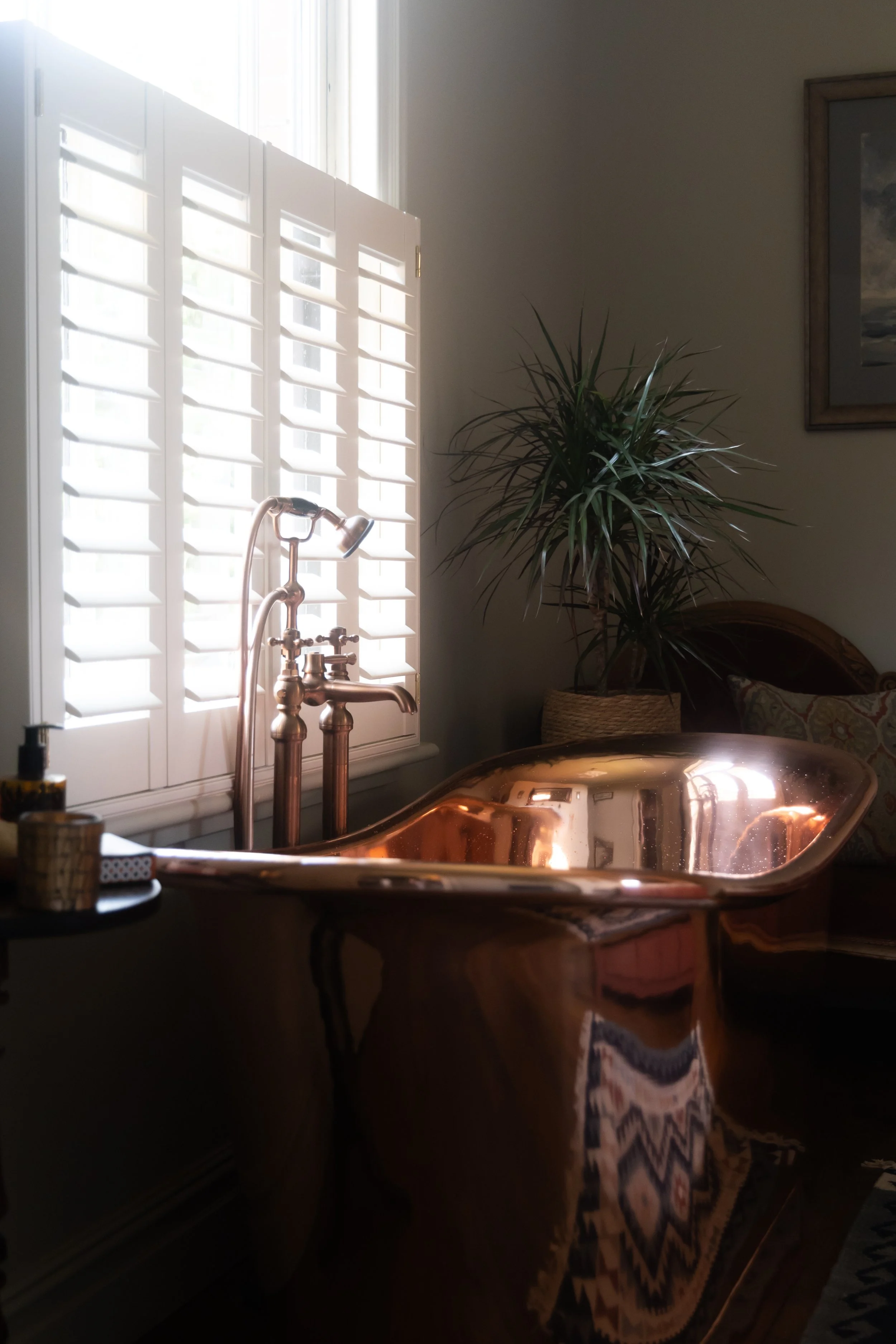 A room with sunlight streaming through white plantation shutters, a brass bathtub, a potted plant next to the tub, and a framed picture on the wall.