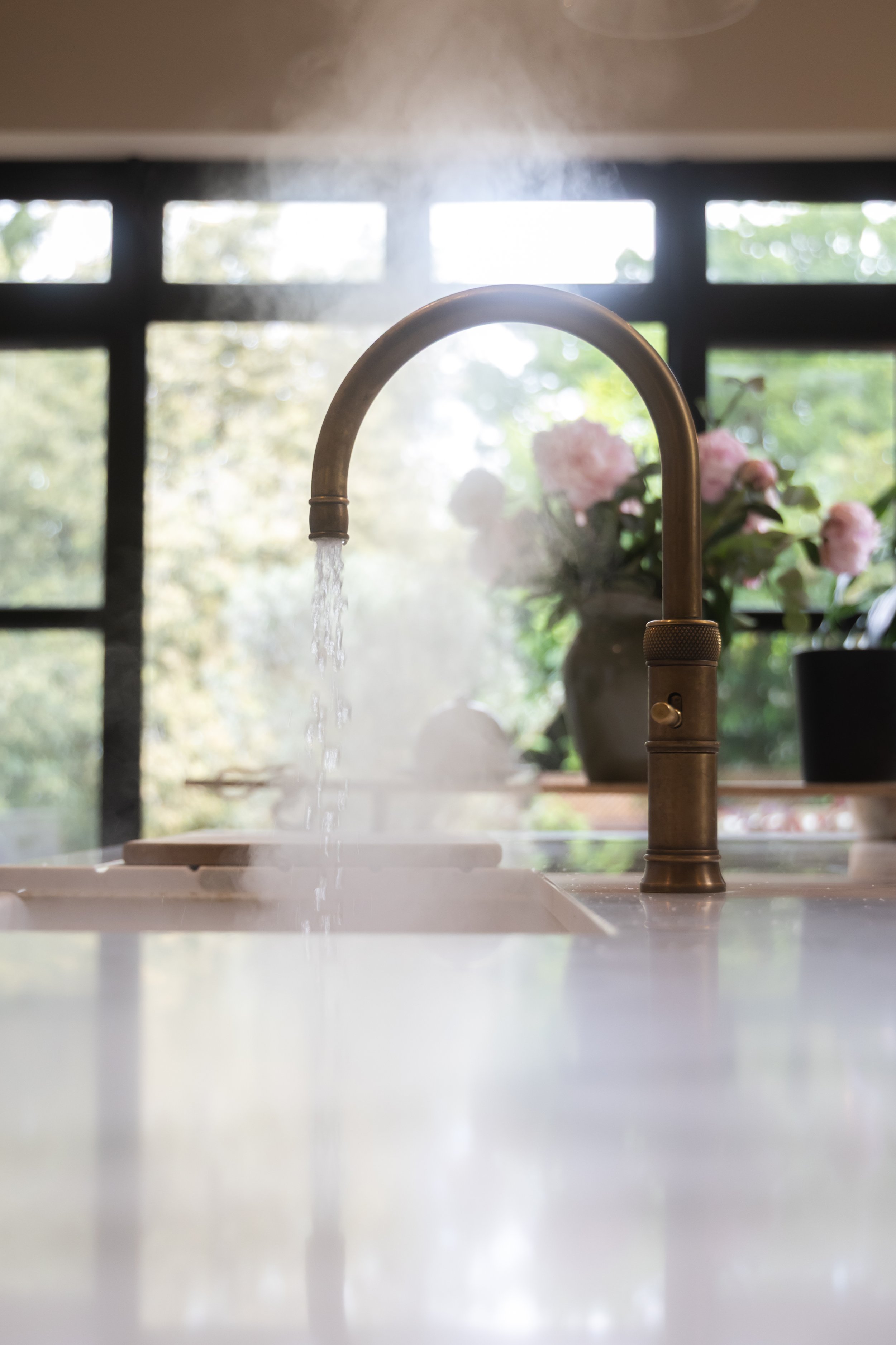 Kitchen sink faucet with water running, steam rising, and a window with greenery in the background.
