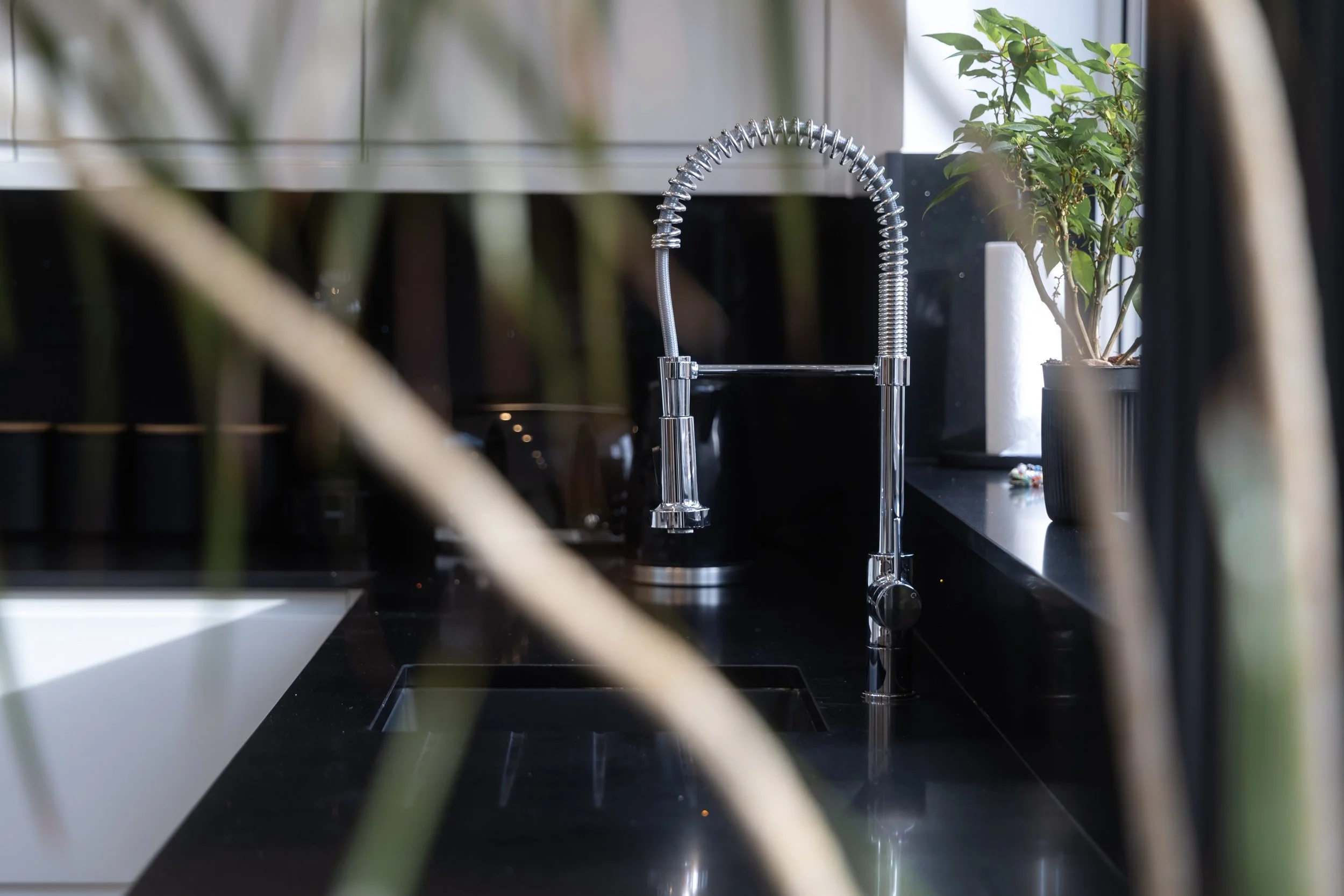 A modern kitchen sink with a chrome flexible faucet on a black countertop, with a potted plant and paper towel roll in the background.