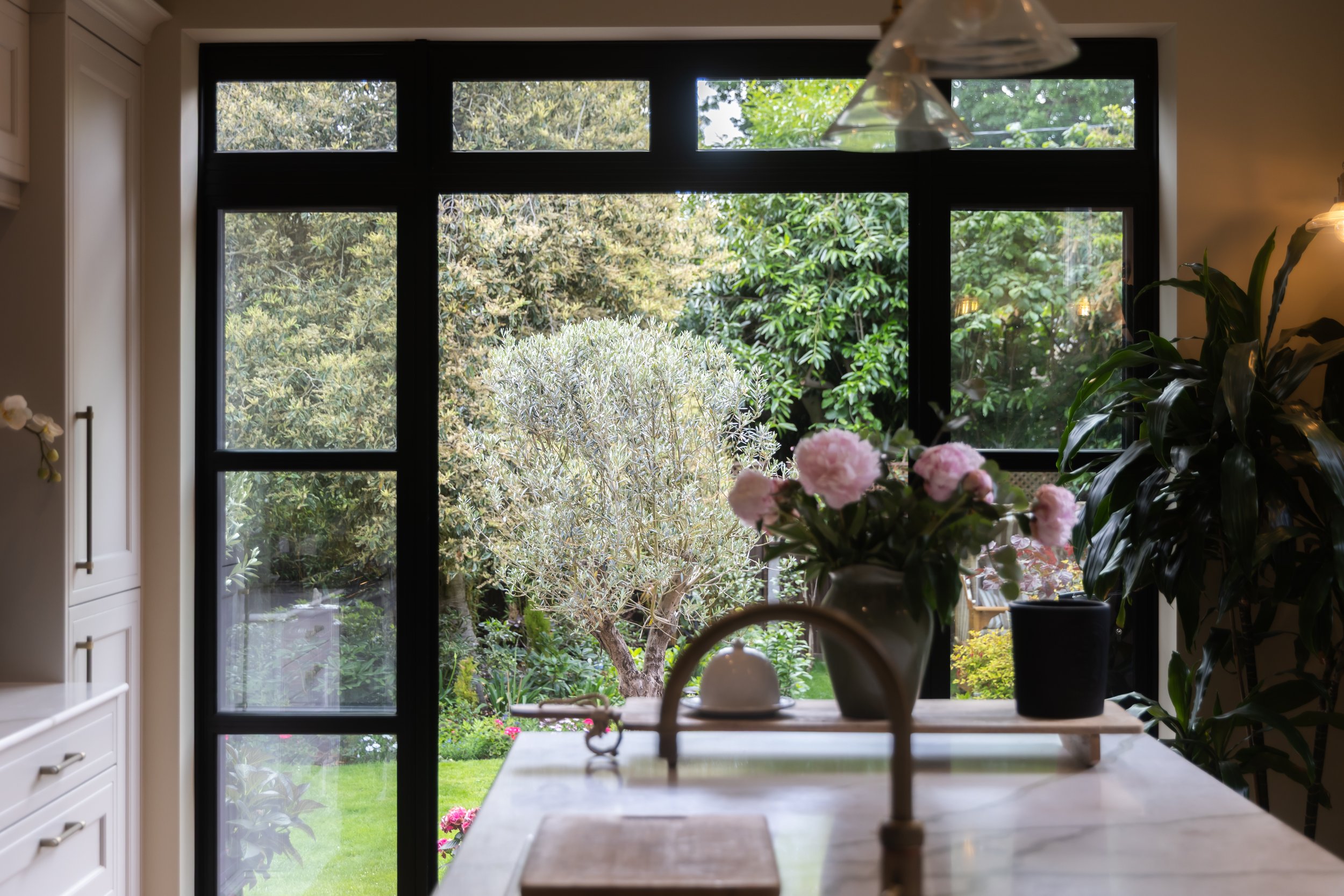 View of a garden outside a large window with black framing, showing trees and greenery. Inside, there are potted plants, pink flowers, and a table with decorative items, including a white vase and a small sculpture.