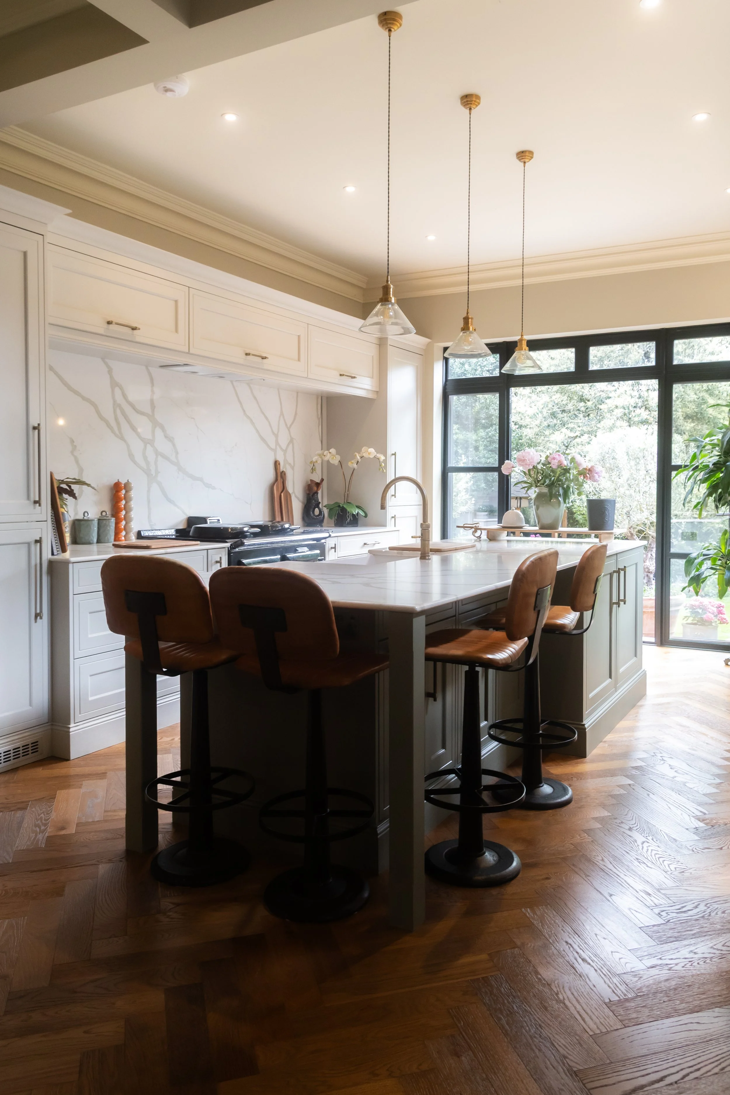 A modern kitchen featuring white cabinets, a marble backsplash, a large island with bar stools, pendant lighting, large windows letting in natural light, and some decorative items including flowers and wooden utensils.