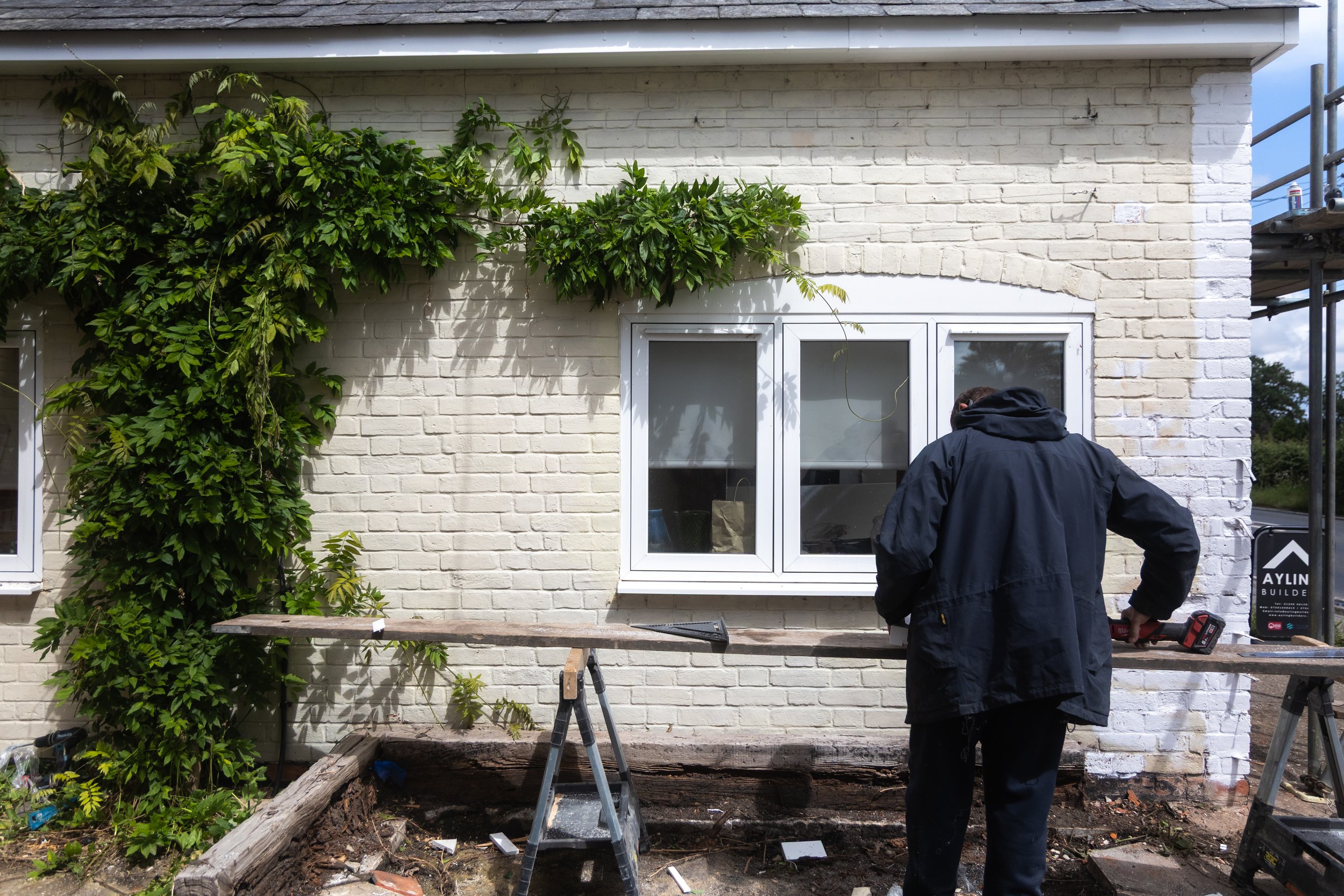 A man working on a house exterior, installing a white window on a white brick wall, with some greenery and scaffolding nearby.