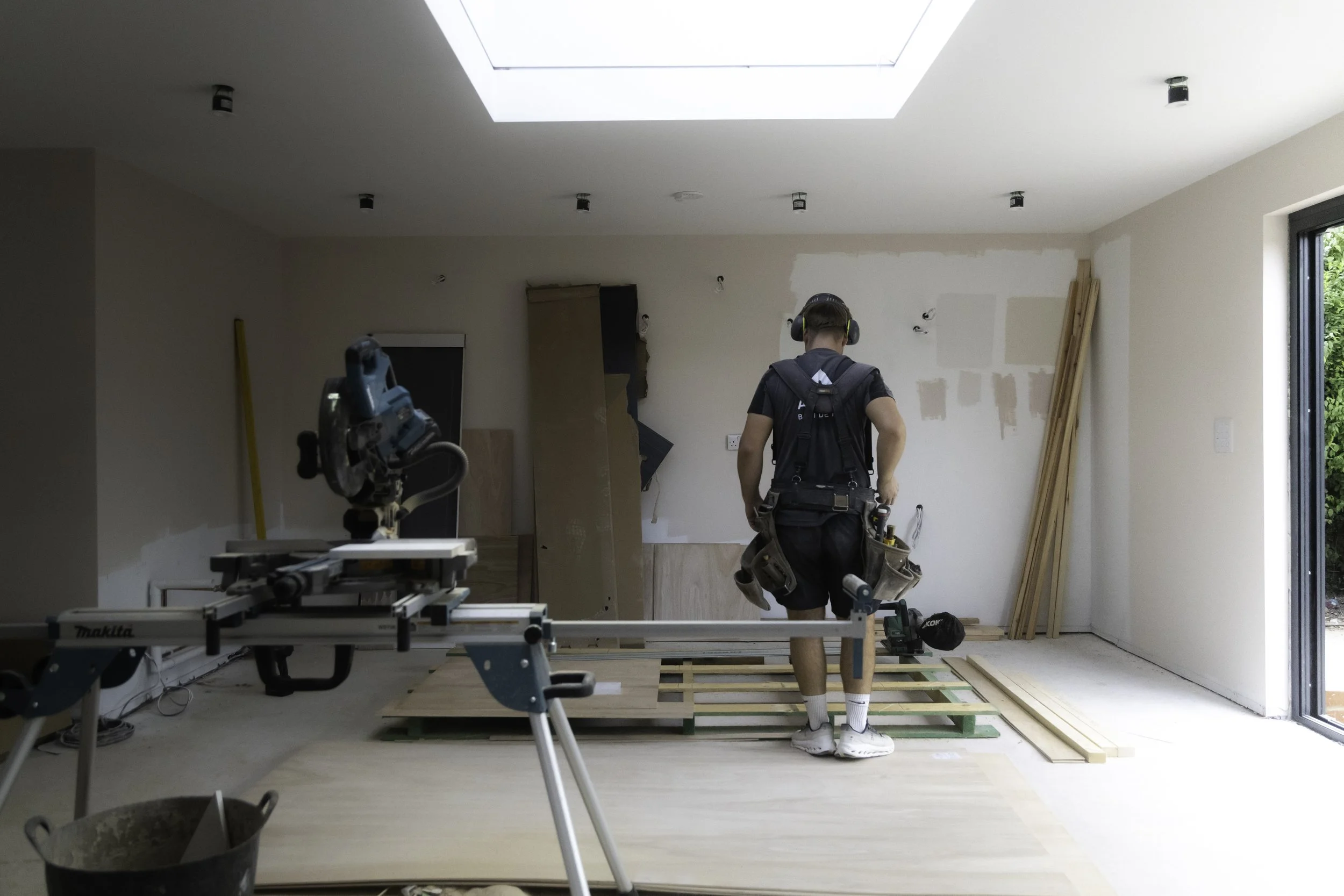 A person working on home renovation in a partially constructed room with wooden flooring and drywall. The person is wearing headphones, a black T-shirt, shorts, and sneakers, and is equipped with a tool belt. Construction tools and materials are scat
