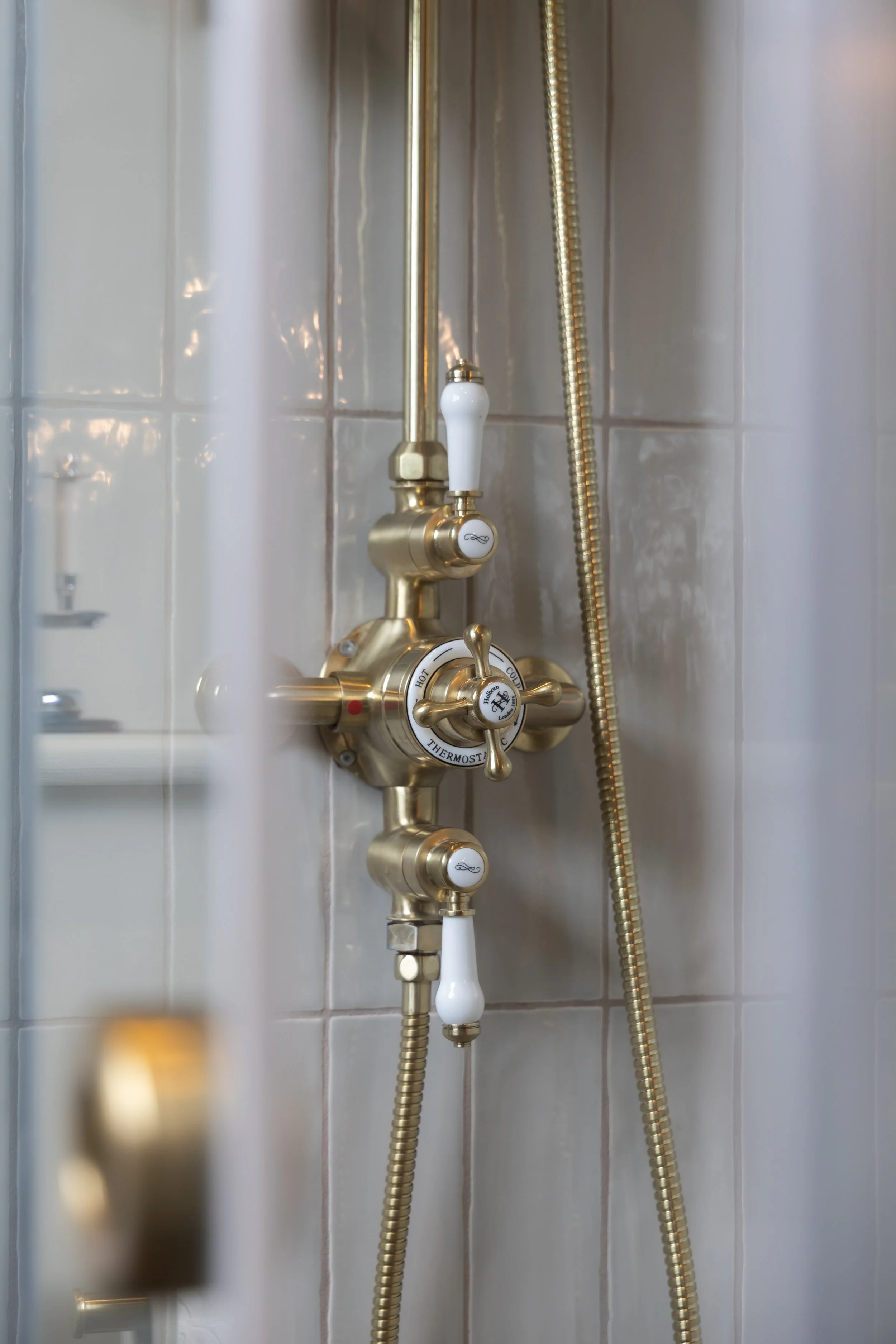 A close-up of a vintage-style shower valve with white handles and gold-colored fixtures, mounted on a tiled bathroom wall, with a flexible gold shower hose.