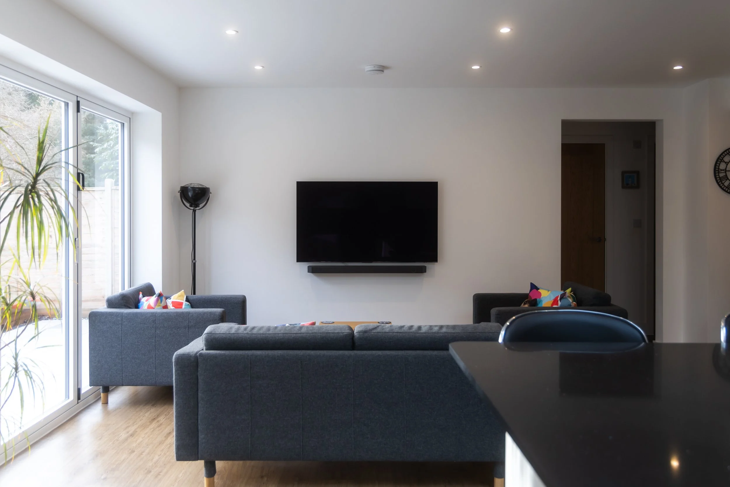 Living room with sliding glass door, gray sofas with colorful pillows, wall-mounted TV, floor lamp, and hallway with a wall clock.