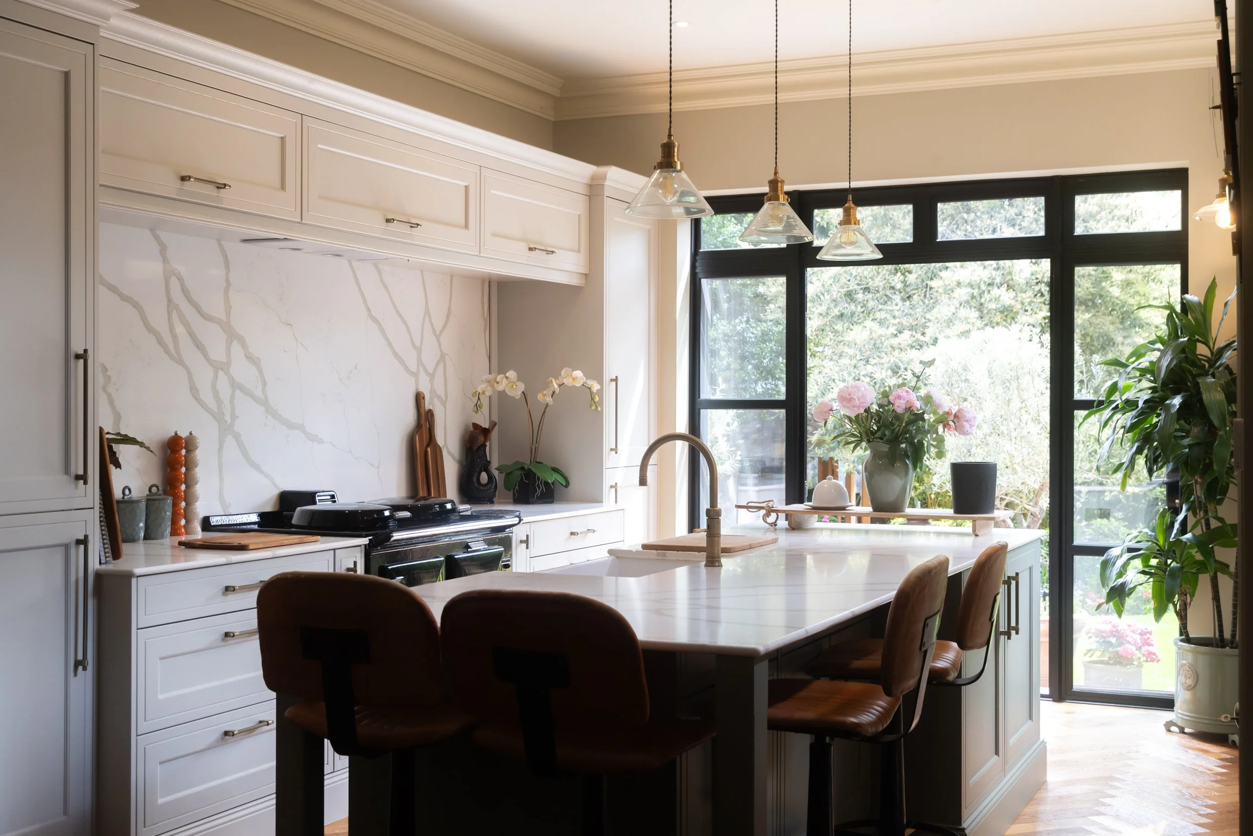 A modern kitchen with white cabinets, a marble backsplash, and large black-framed windows letting in natural light. There are hanging pendant lights above the kitchen island and various plants and decor on the counters and windowsill.