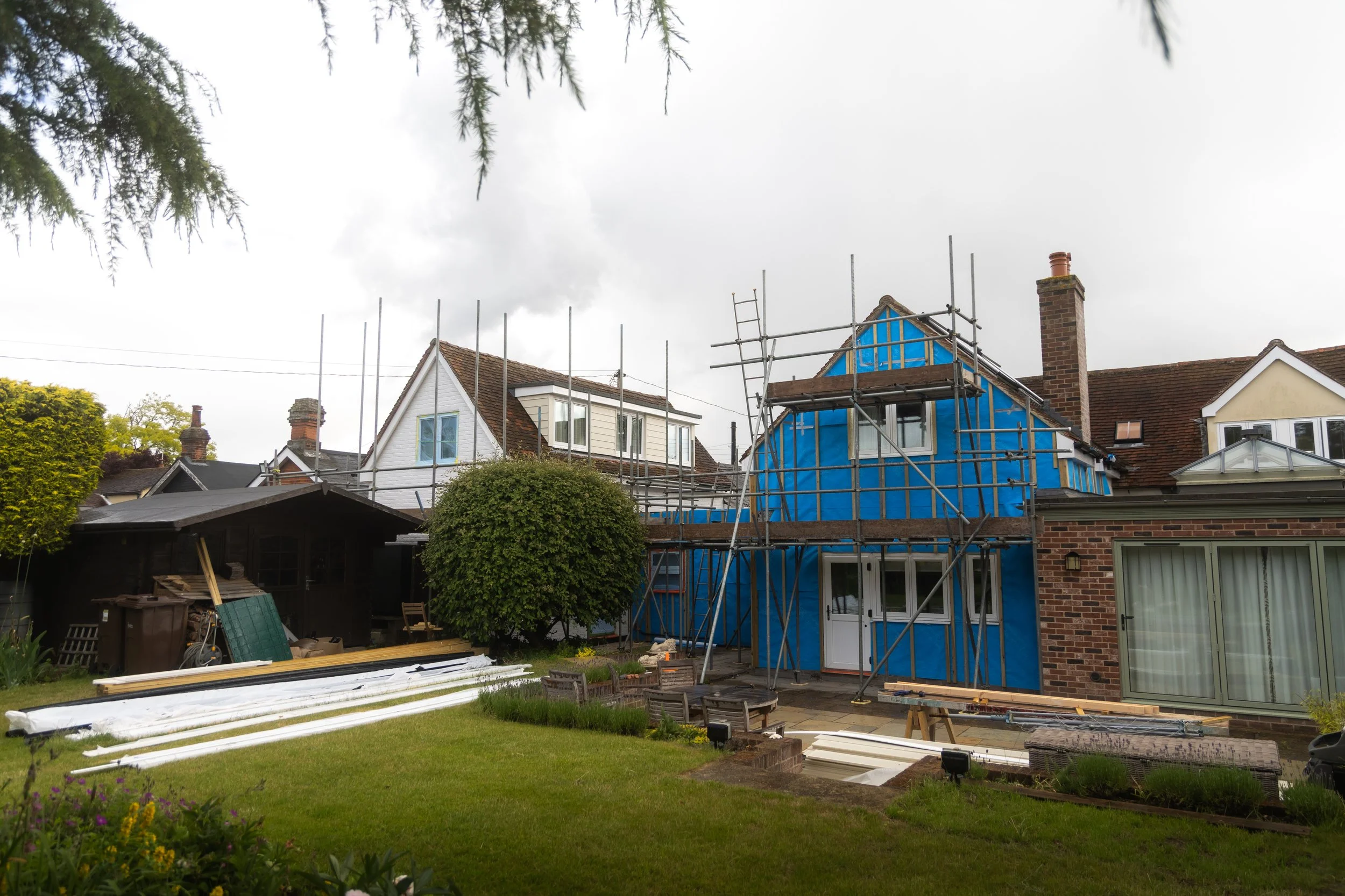 A house under renovation with scaffolding on the front and blue protective sheeting, surrounded by trees and a garden with flowers and outdoor furniture.