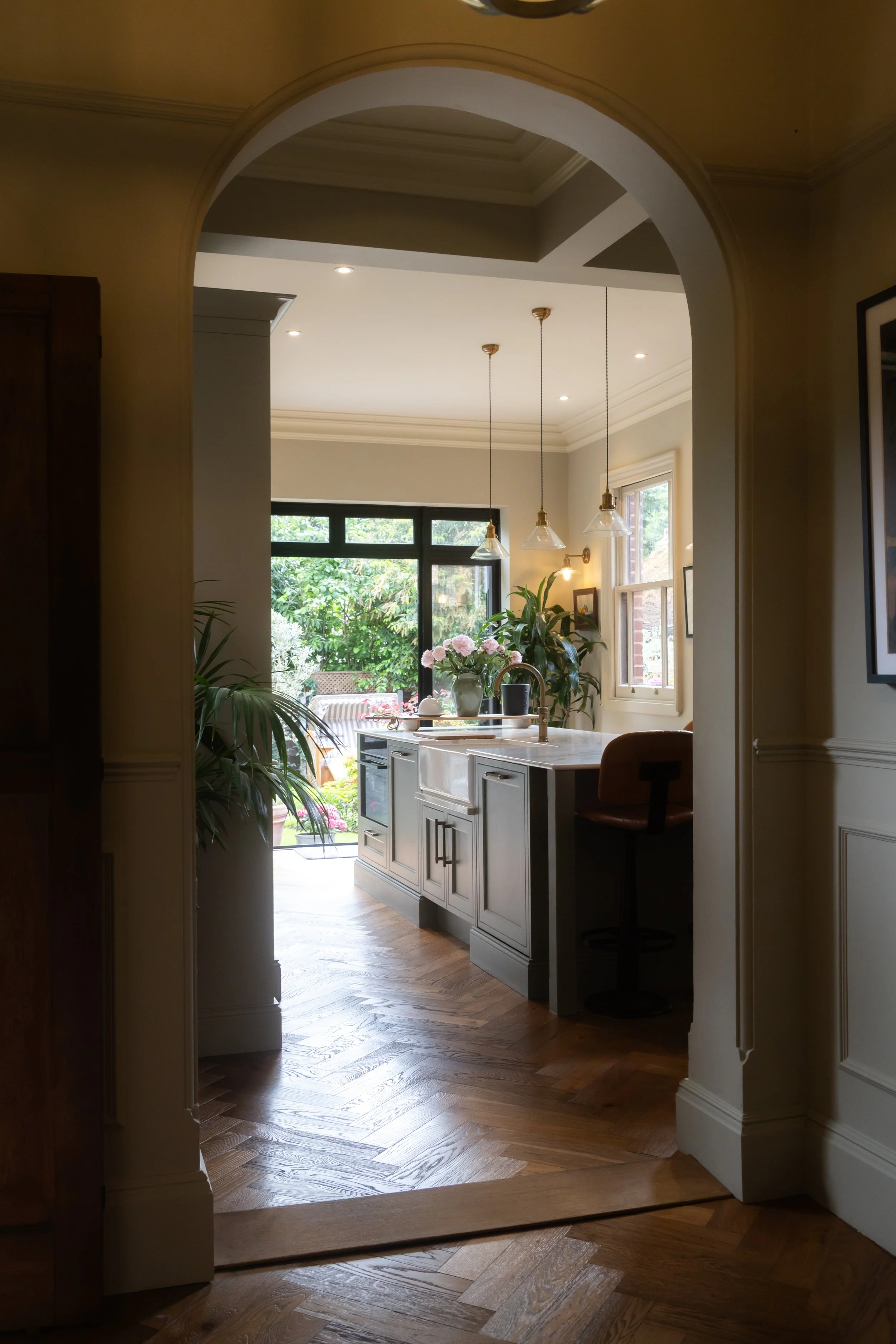 View of a bright, modern kitchen from an arched doorway, with a kitchen island, pendant lights, large windows, and lush greenery outside.