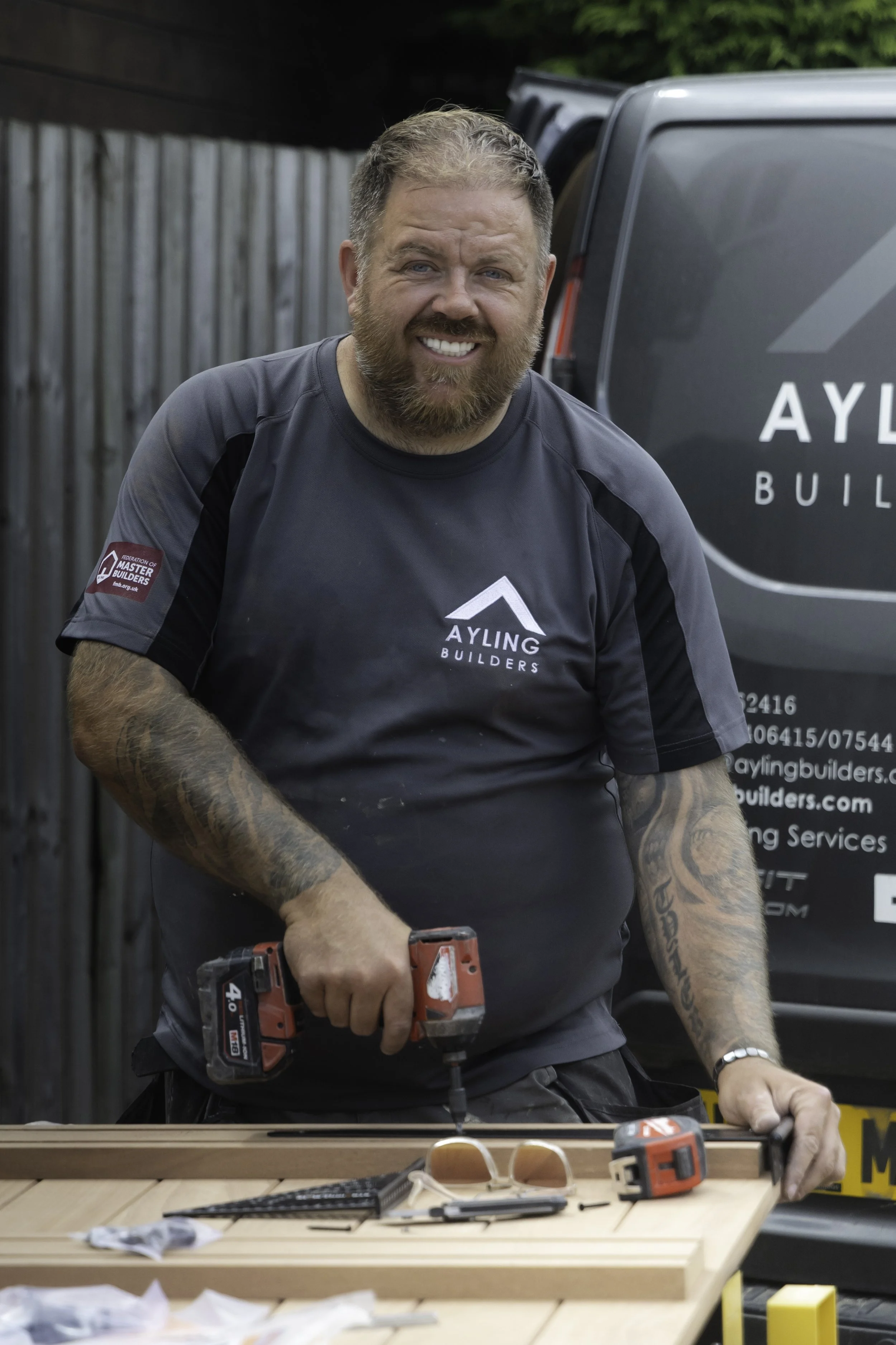 Man with a beard and tattoos using a cordless drill at a workbench outdoors, with tools and sunglasses on the table, and a black van in the background.