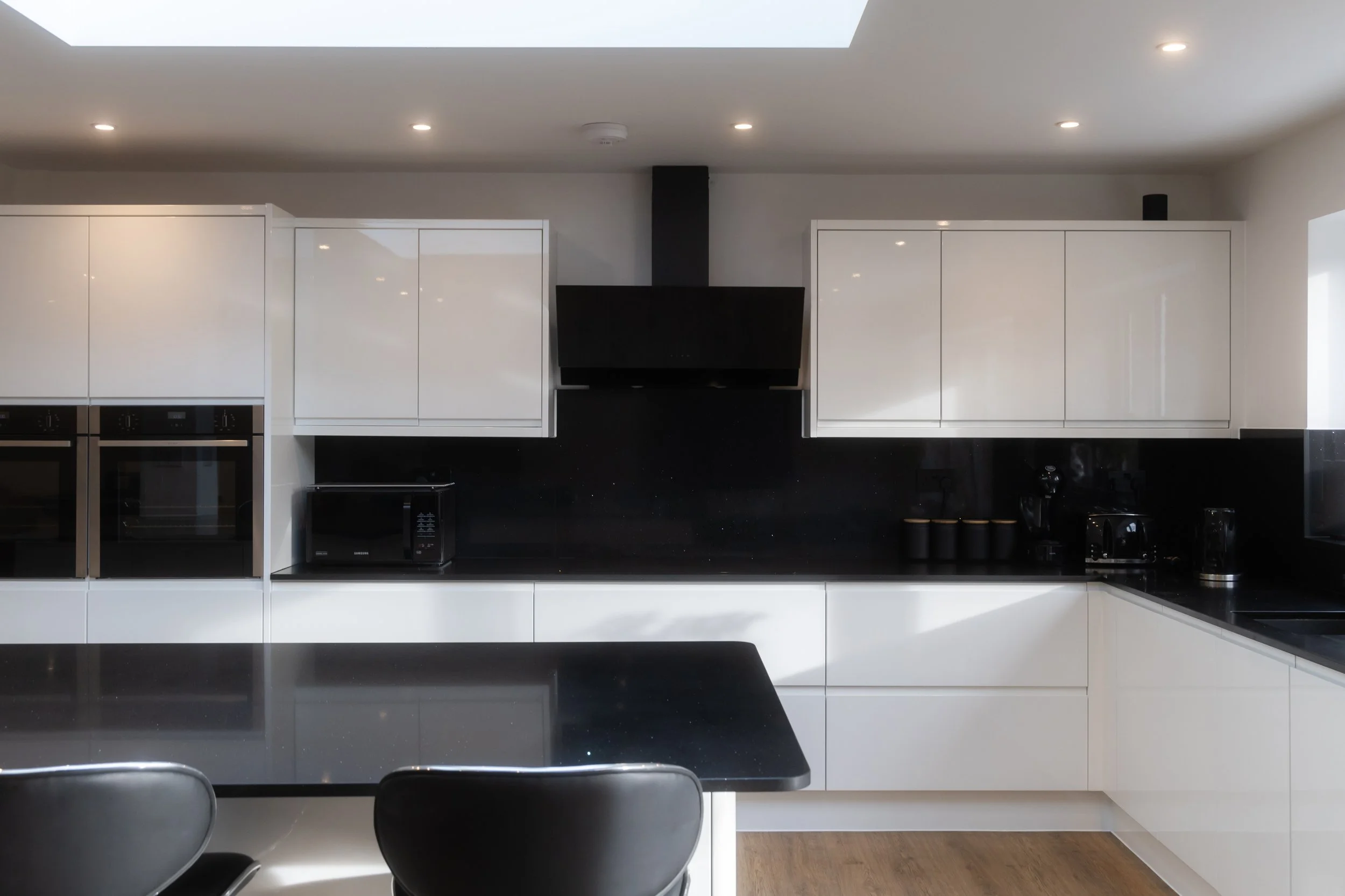 Modern kitchen with white cabinets, black countertop, built-in oven, microwave, and black range hood. A ceiling skylight and recessed lighting are visible. There is a black kitchen island or table with black chairs in the foreground.