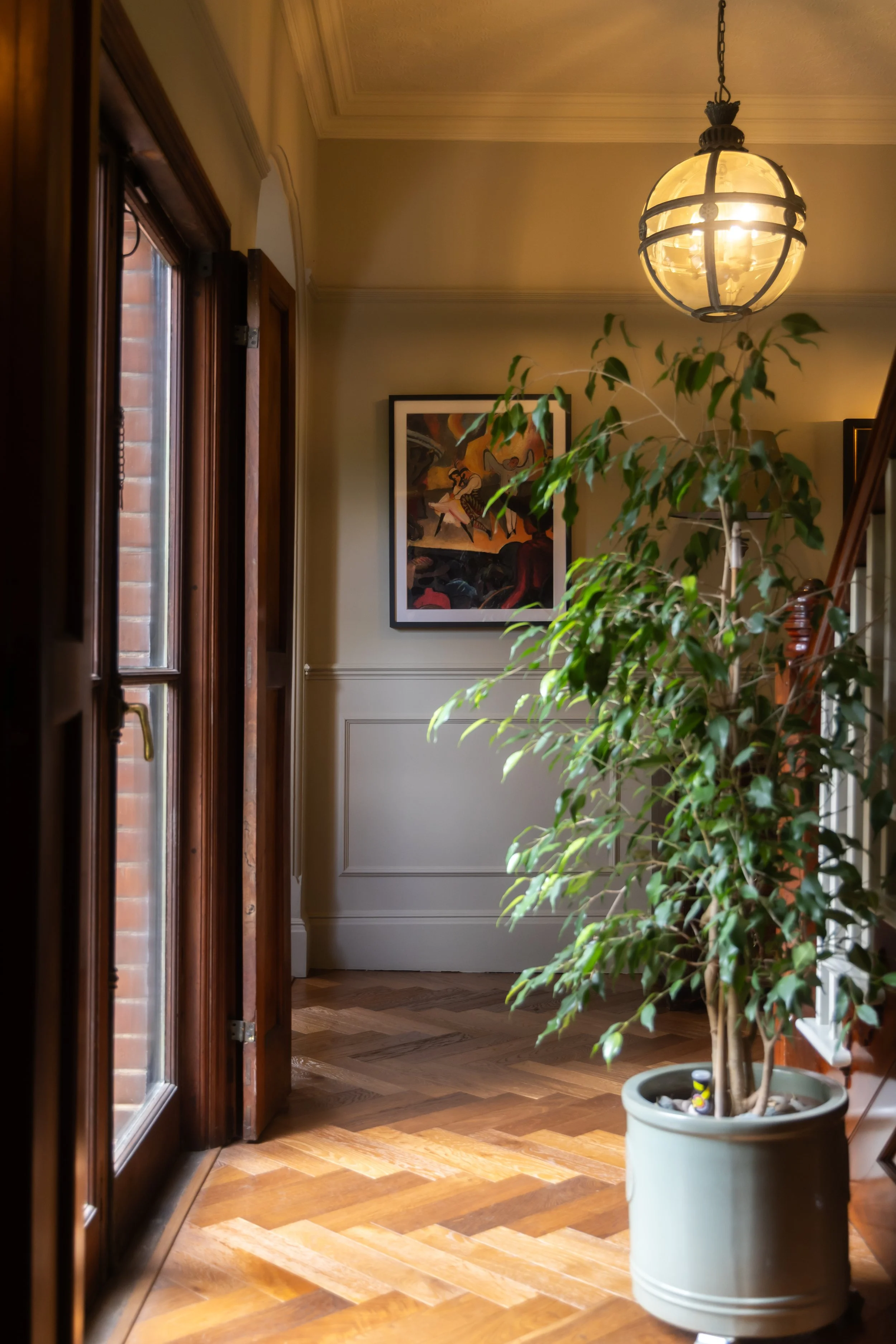 Interior of a home hallway with wooden floors, glass-paneled door, framed artwork on the wall, potted plant with large green leaves, and a hanging ceiling light.