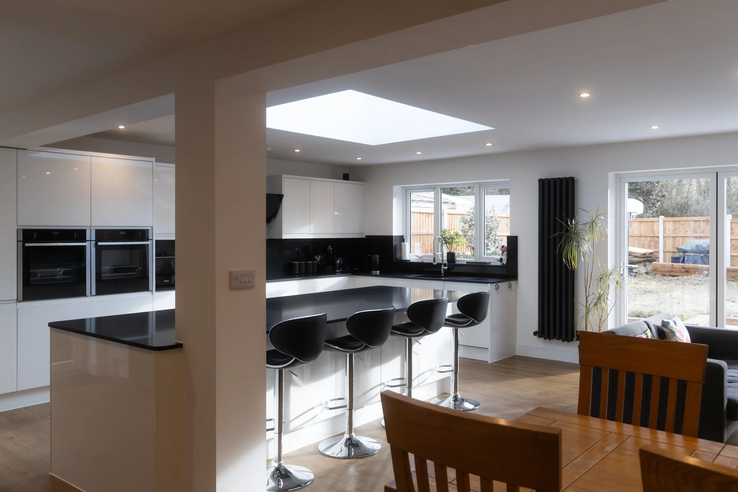 Modern kitchen with white cabinets, black countertops, and bar seating with four black chairs, large window with black curtains, and sliding glass door leading to outdoor backyard.