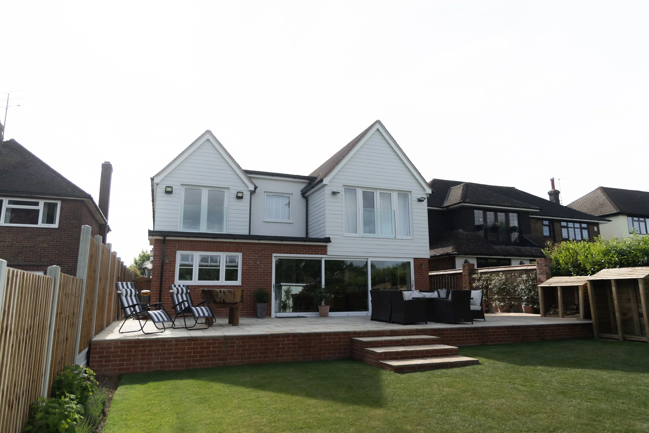 Backyard patio with chairs and a view of a white house with large windows, brick base, and a lawn.